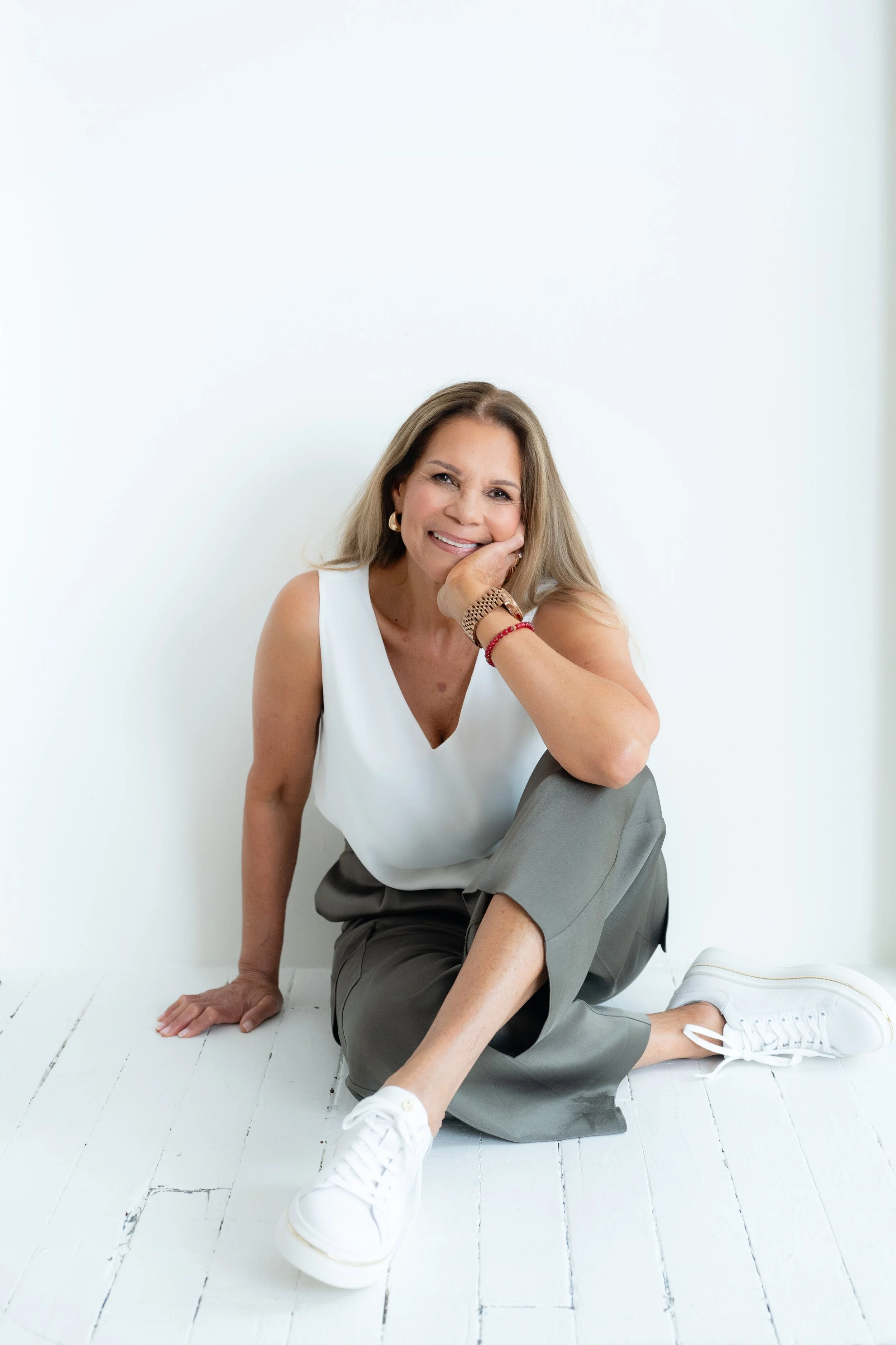 Woman sitting on a white wooden floor against a white wall, smiling, in casual clothing.