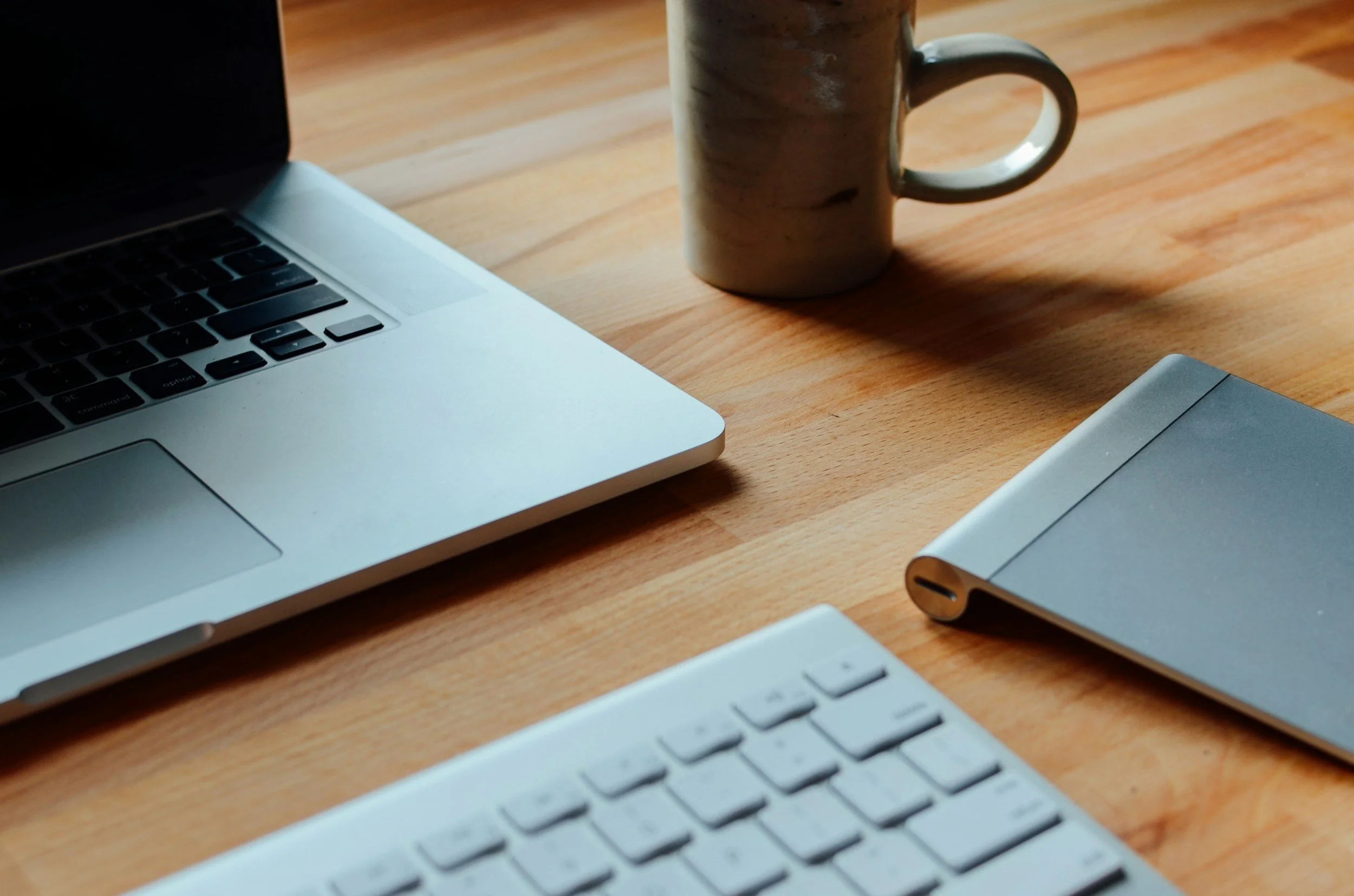 A wooden desk with a silver laptop on the left, a white ceramic mug in the middle, and part of a white keyboard and a silver device on the right.