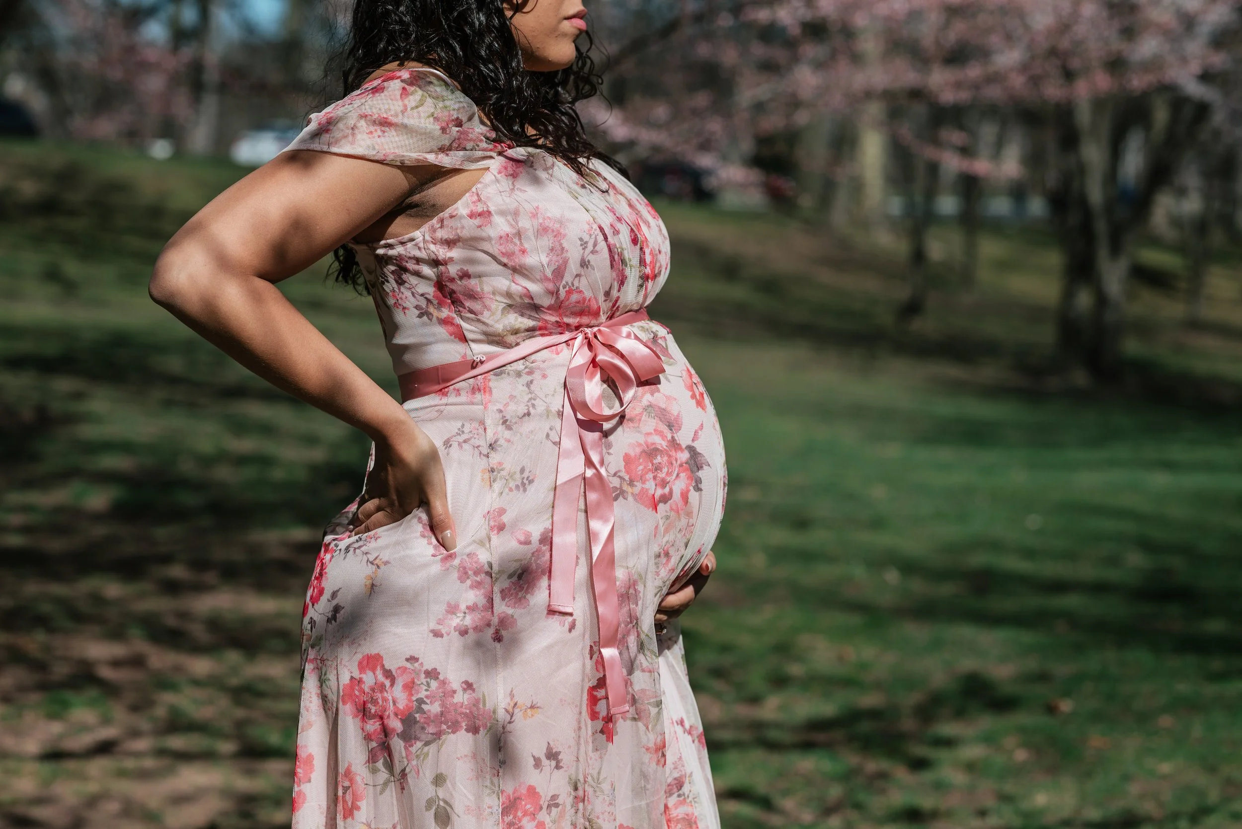 Side view of a pregnant woman wearing a pink floral dress with a satin ribbon tied around her waist, standing outdoors in a park with trees and grass in the background.