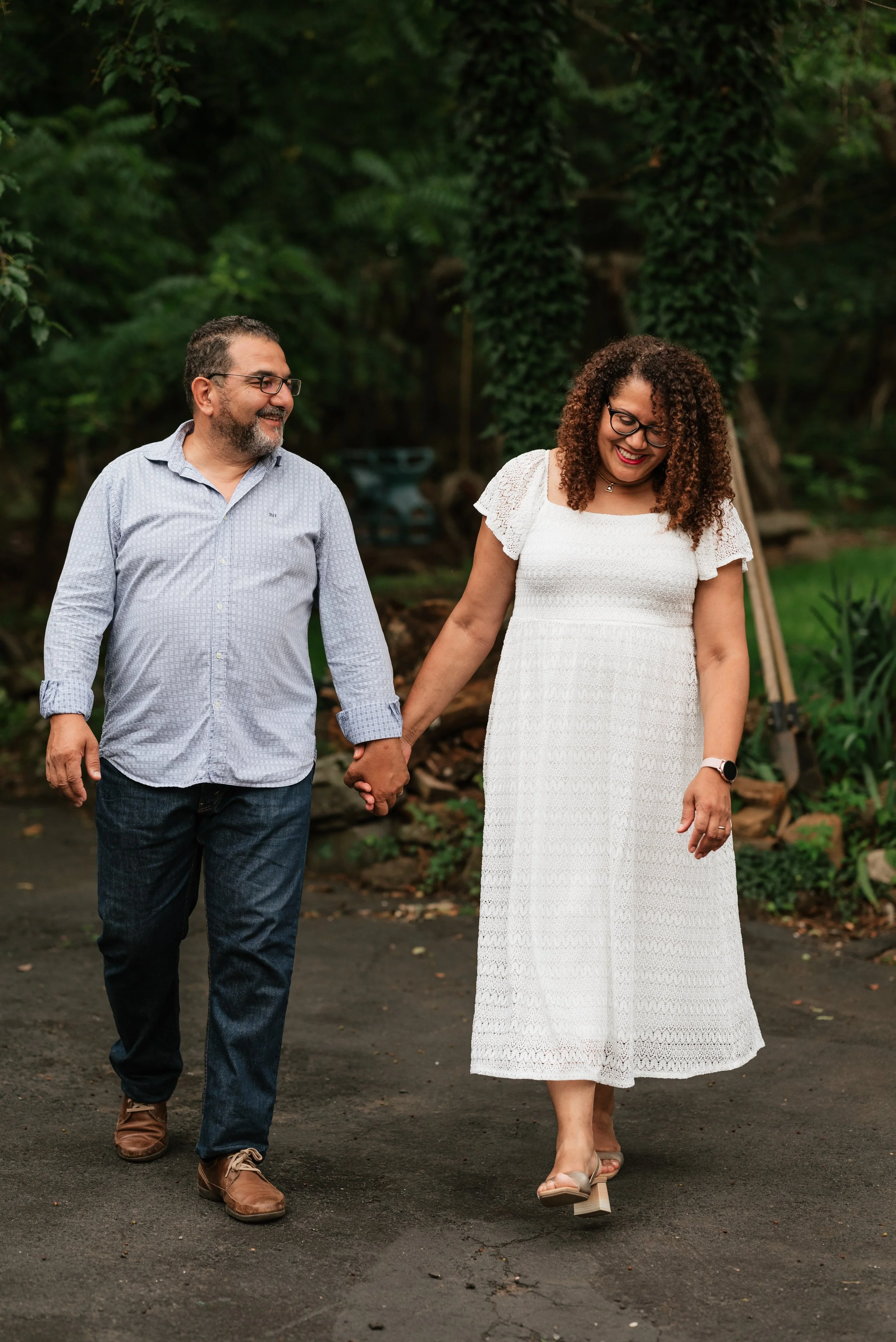 A couple walking hand in hand outdoors in a lush green garden, smiling and looking at each other.