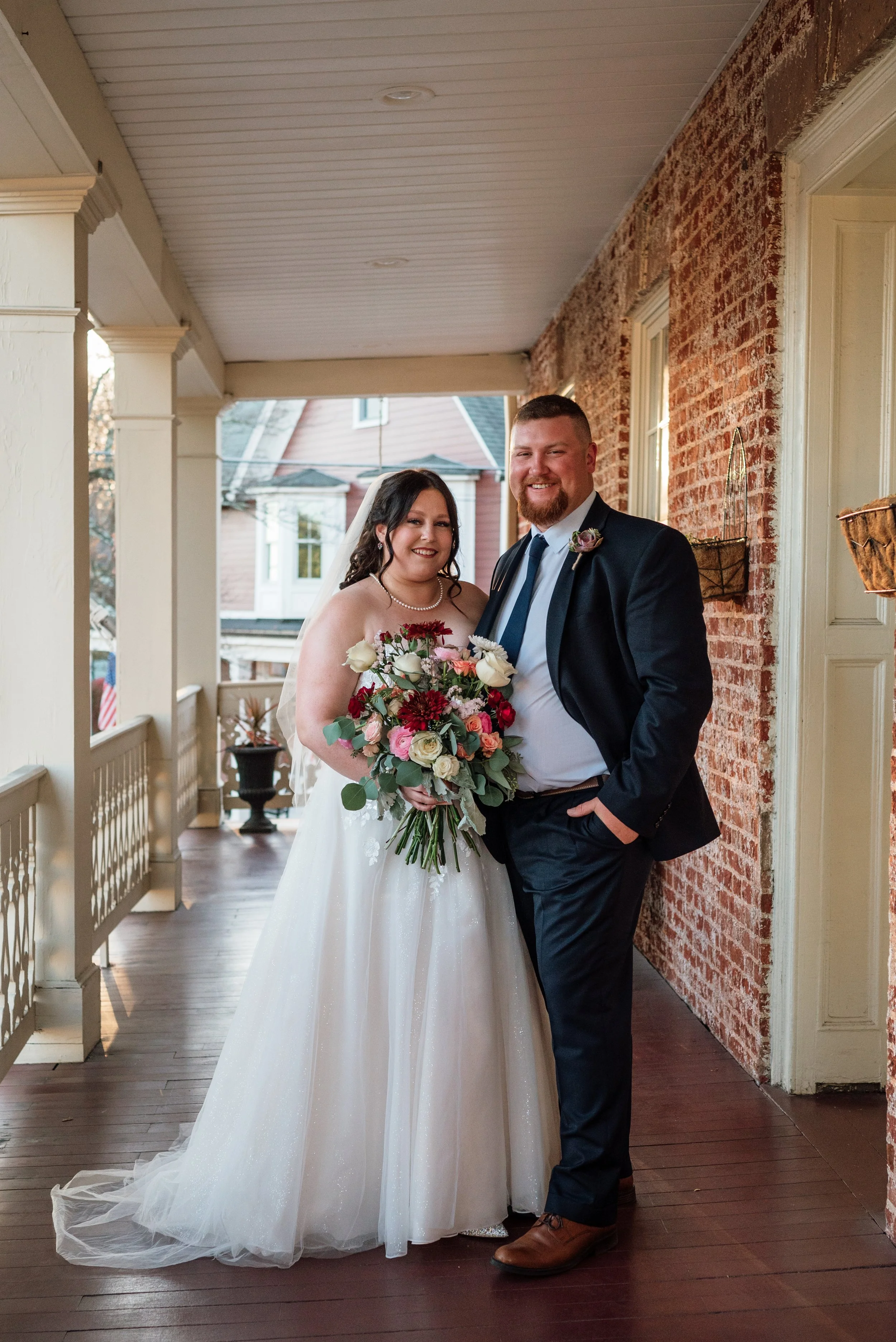 A bride and groom standing together on a porch, smiling, with the bride holding a bouquet of flowers.