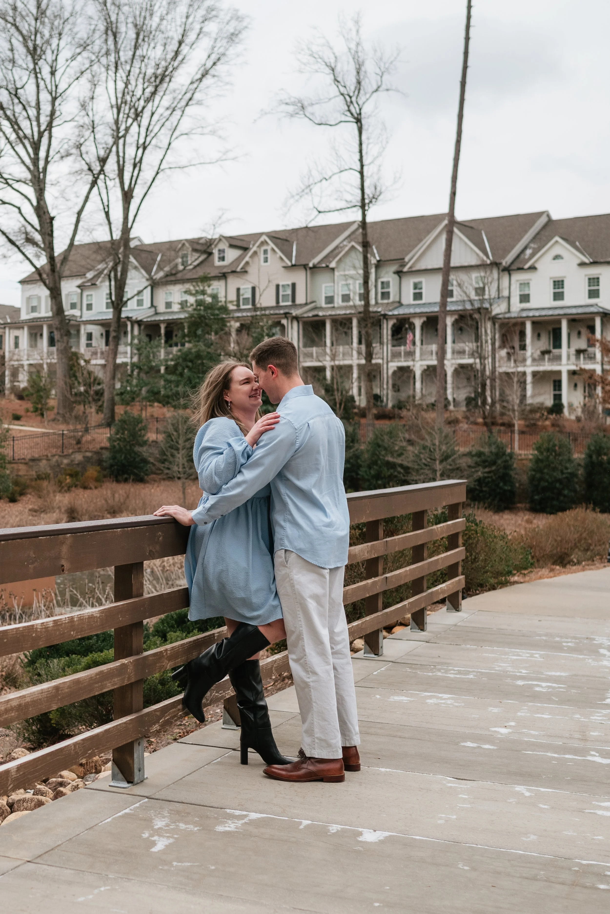 A couple embracing and smiling on a bridge with trees and a house in the background during their engagement photoshoot in Alpharetta, GA.