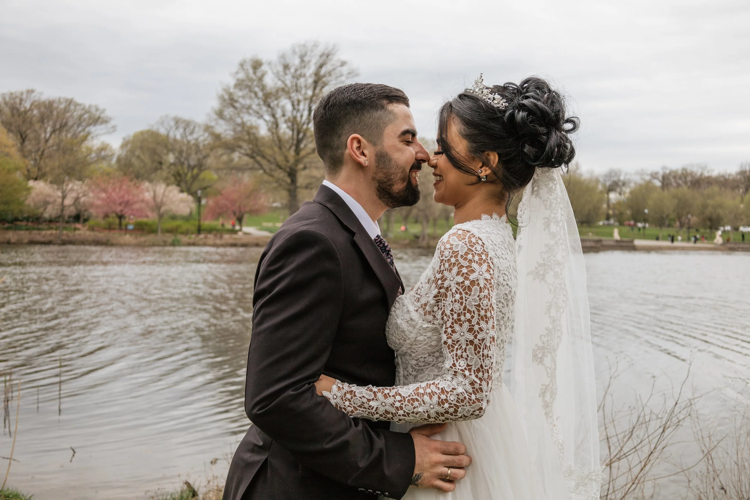 A newlywed couple stands close by a lake, smiling and touching foreheads, with trees and pink blossoms in the background on a cloudy day.