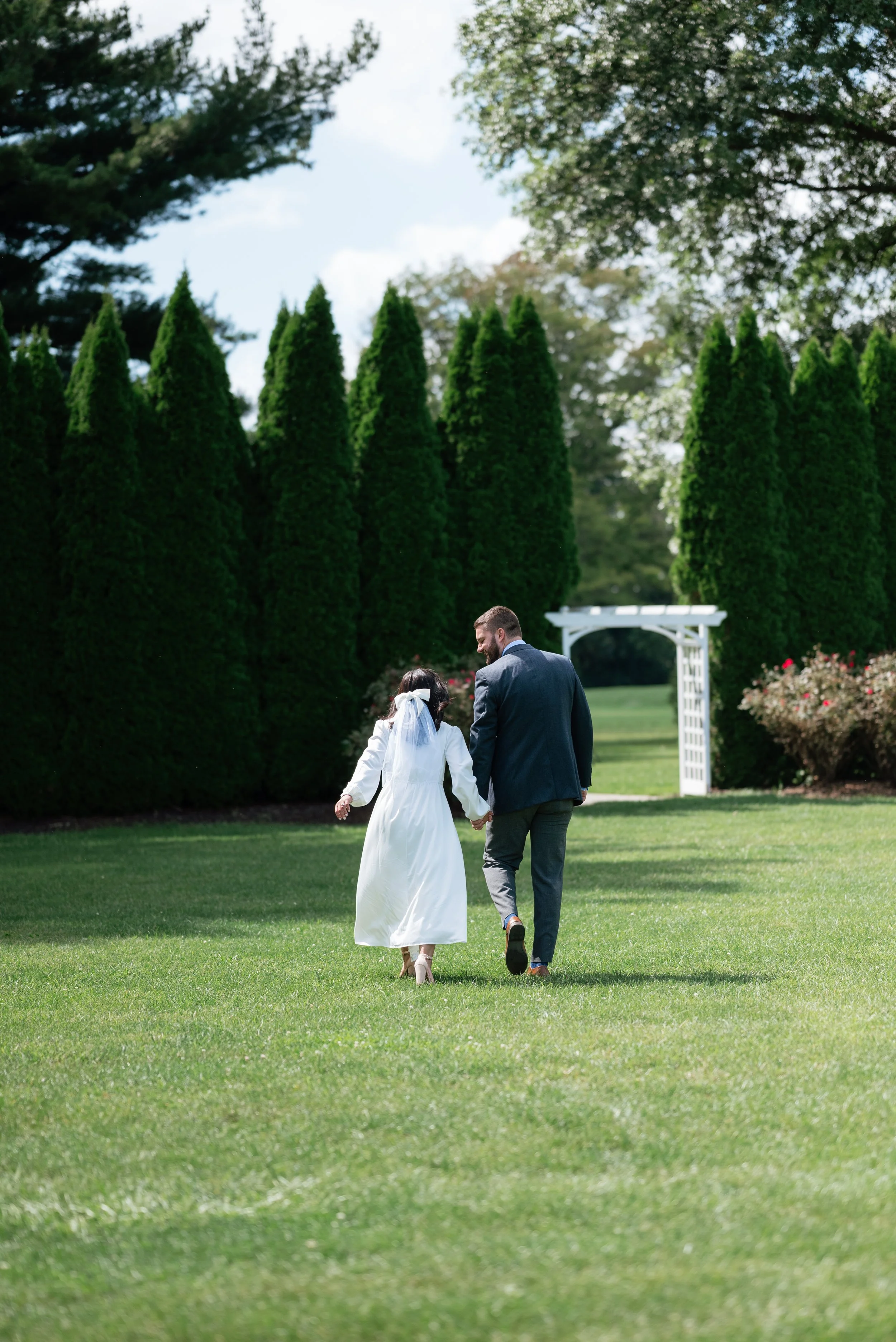 A newlywed couple walking hand-in-hand on a green lawn towards a white arch, with tall trees in the background.