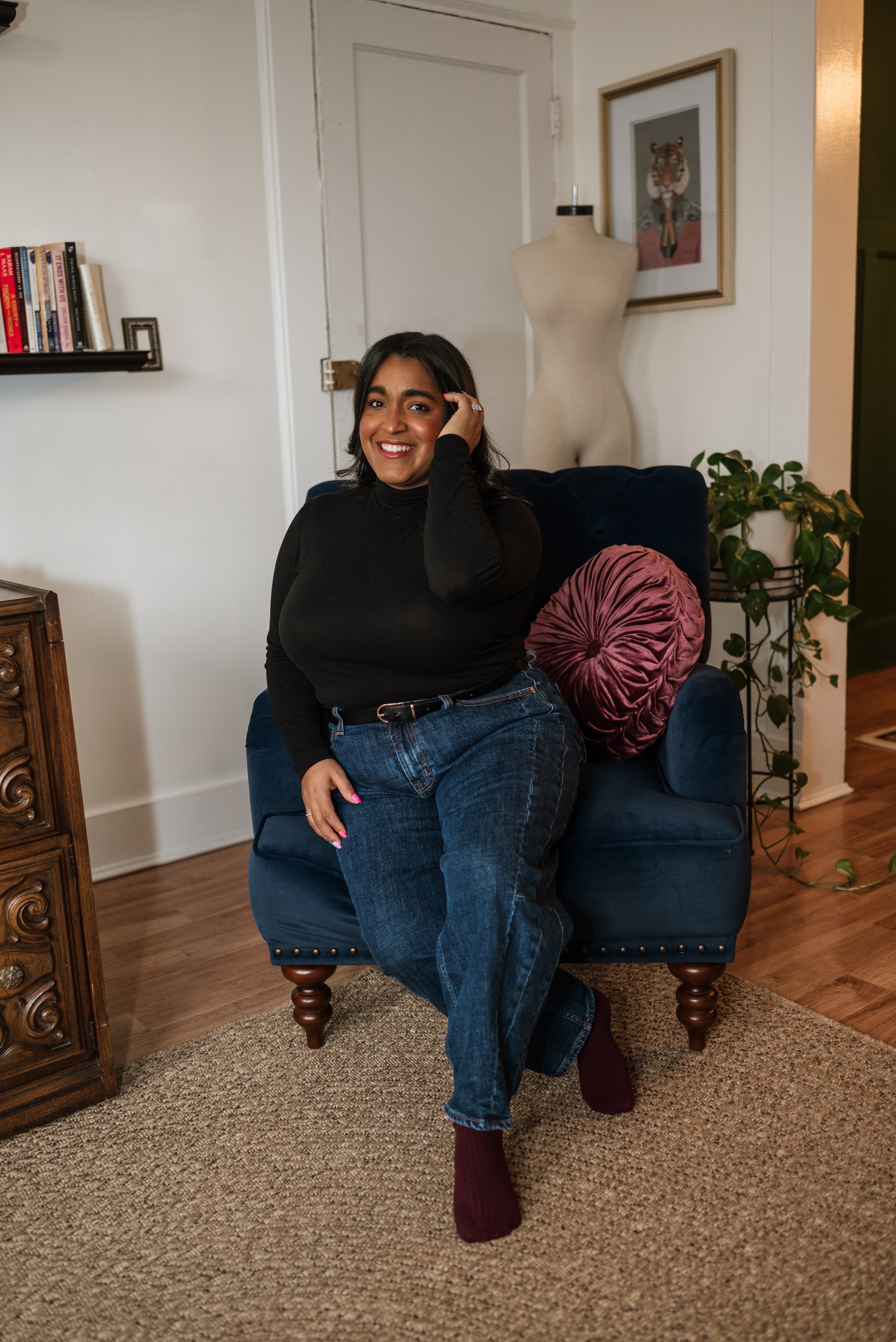 Photographer branding shoot sitting on a blue armchair in a living room, smiling, wearing a black turtleneck and jeans, with a rose-colored pillow behind her, next to a potted plant and wall art.