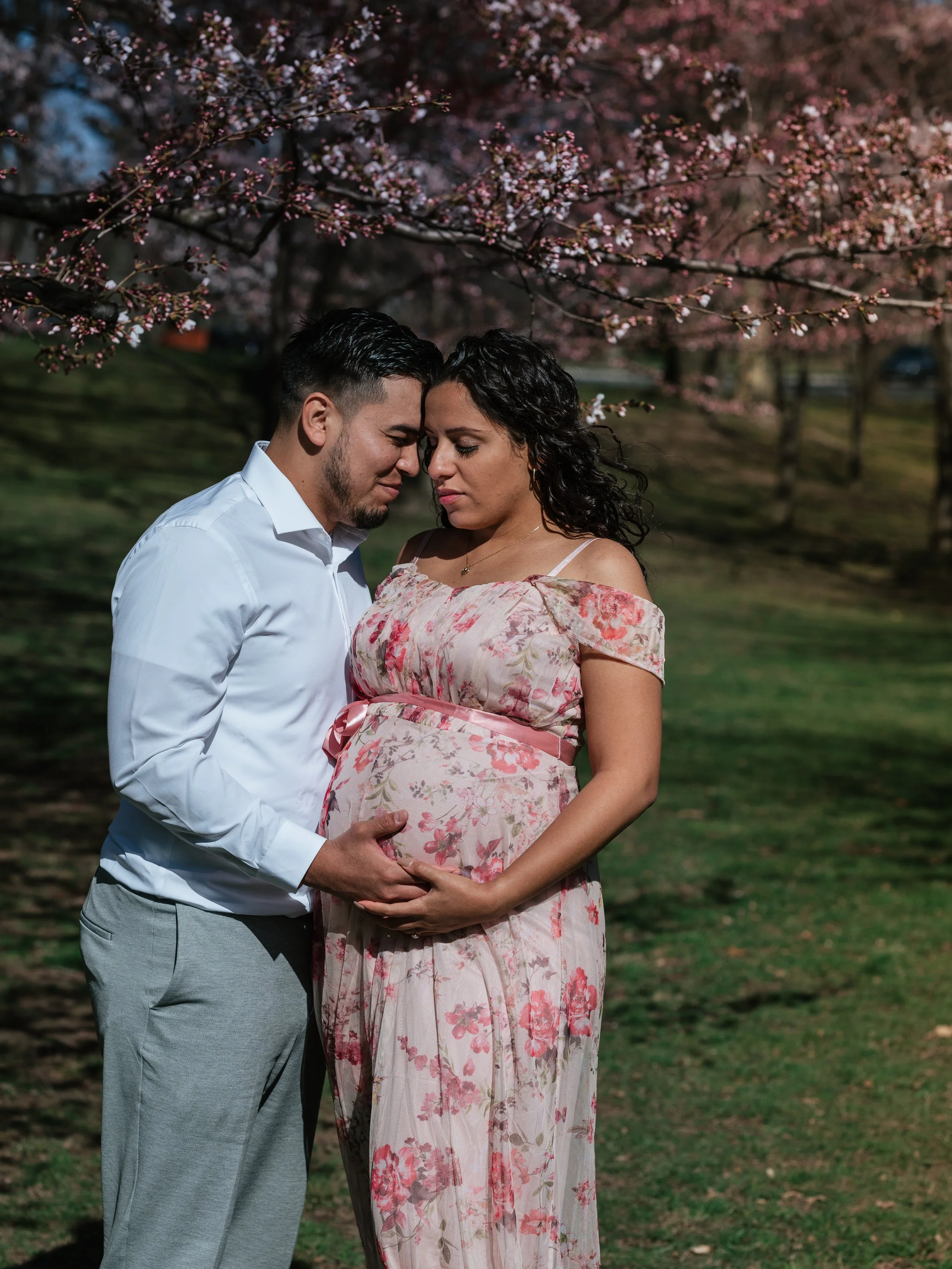 A pregnant woman in a floral dress and a man in a white shirt and gray pants stand close together outdoors under pink flowering trees, their foreheads touching, gently holding her baby bump.