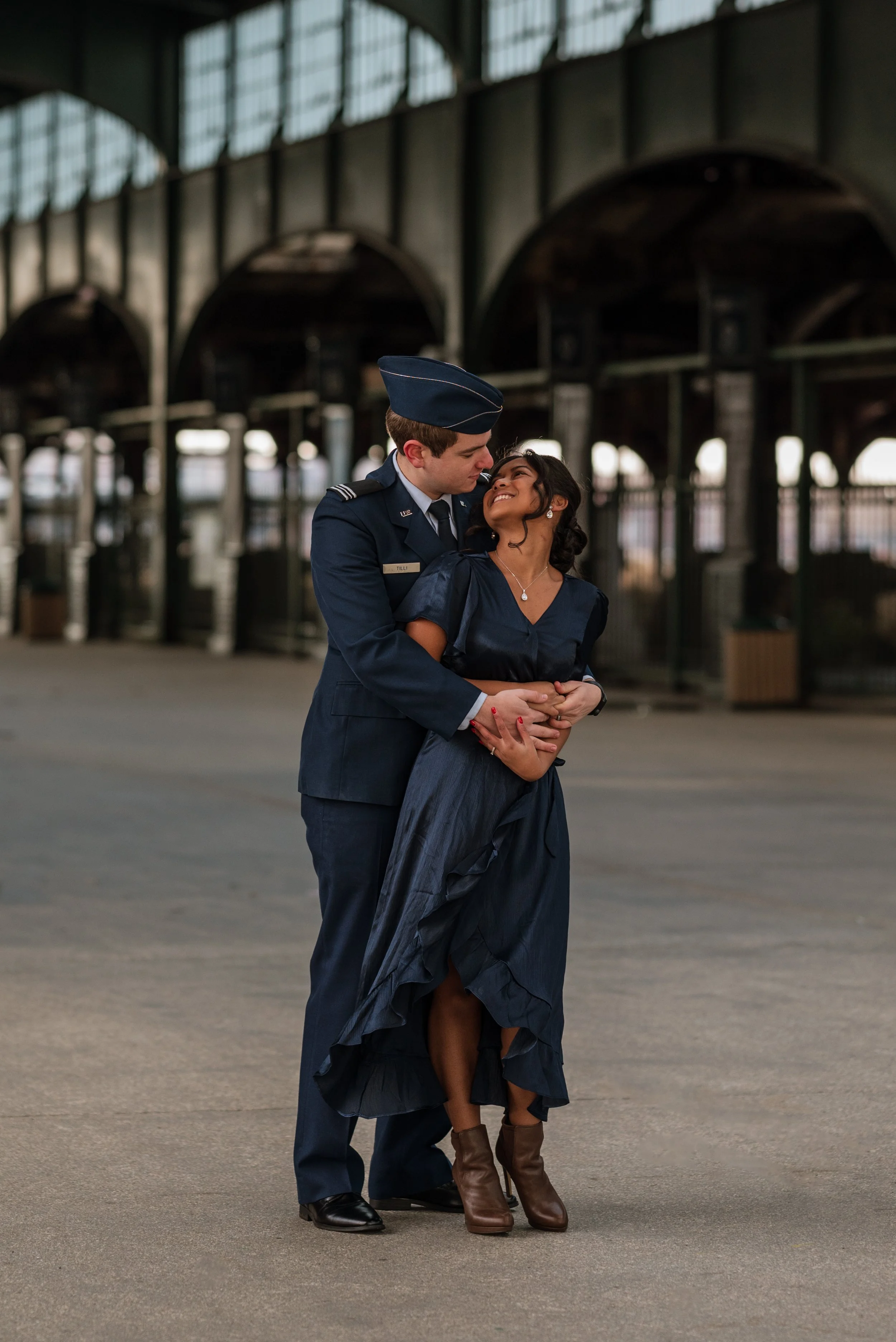 A couple dressed in vintage-style outfits, with the man in a navy blue military uniform and the woman in a navy dress with boots, embracing under a large, industrial-style structure.
