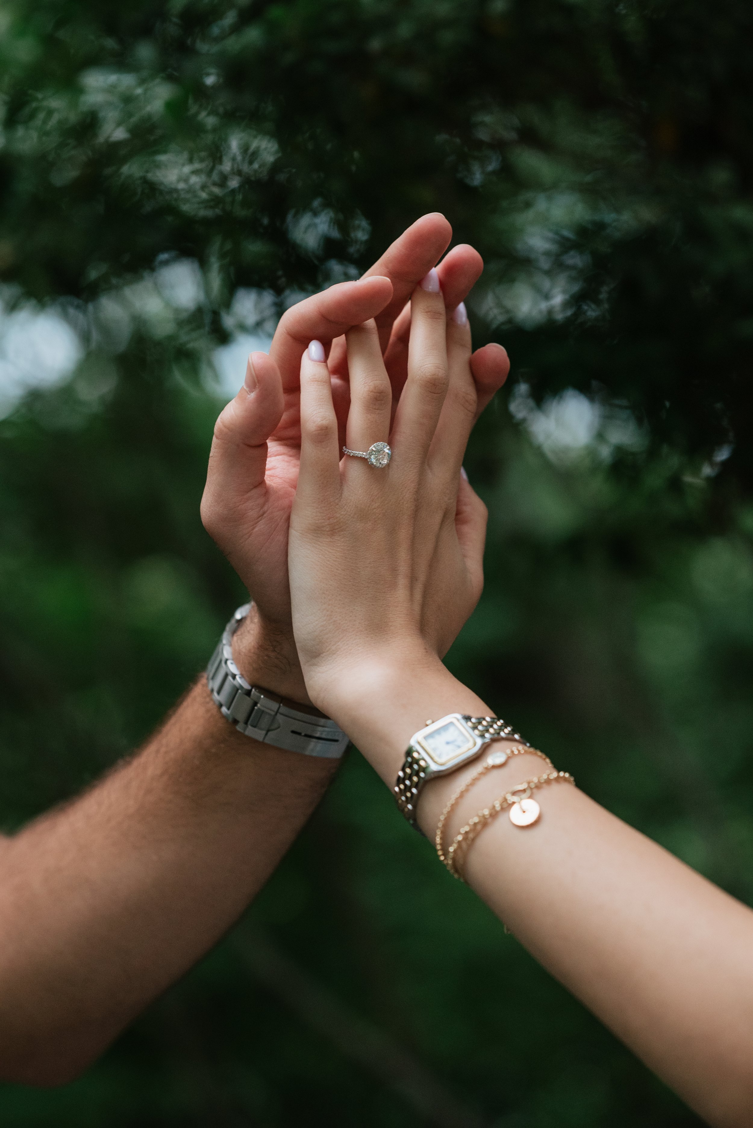 Close-up of a couple's hands with an engagement ring, set against a blurred green forest background.