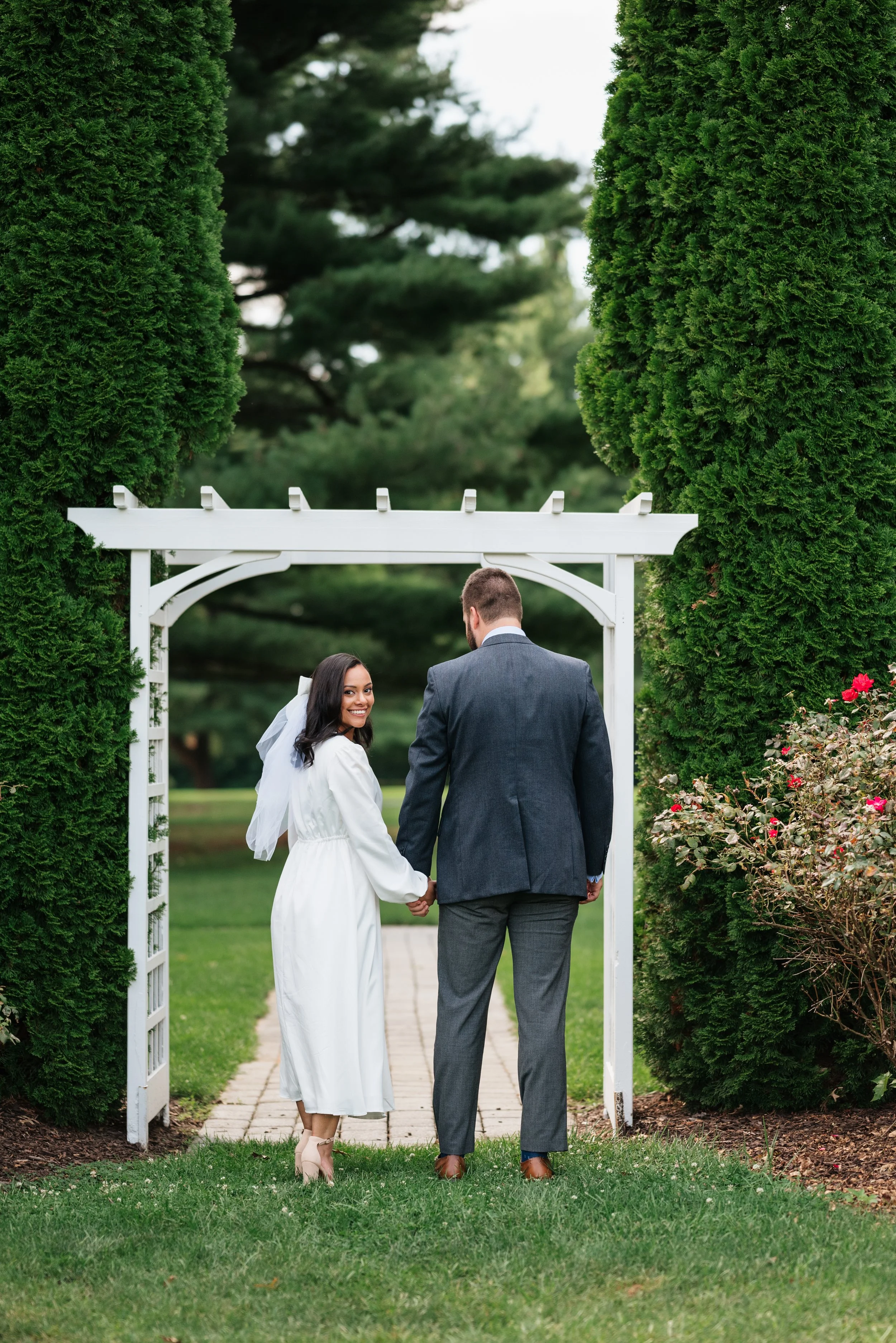 A woman in a white dress holding hands with a man in a grey suit walking under a white garden archway surrounded by green trees and bushes.