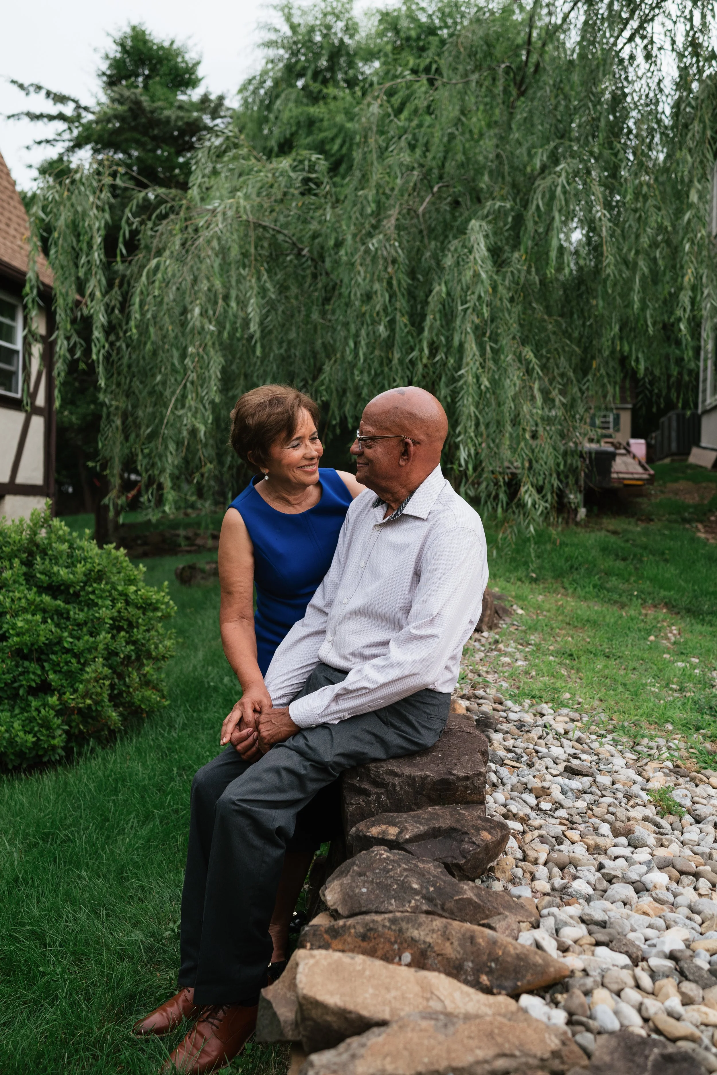 A smiling woman in a blue dress and an elderly man in a white shirt and dark pants sitting on a stone wall outdoors, holding hands, with lush trees and a house in the background.