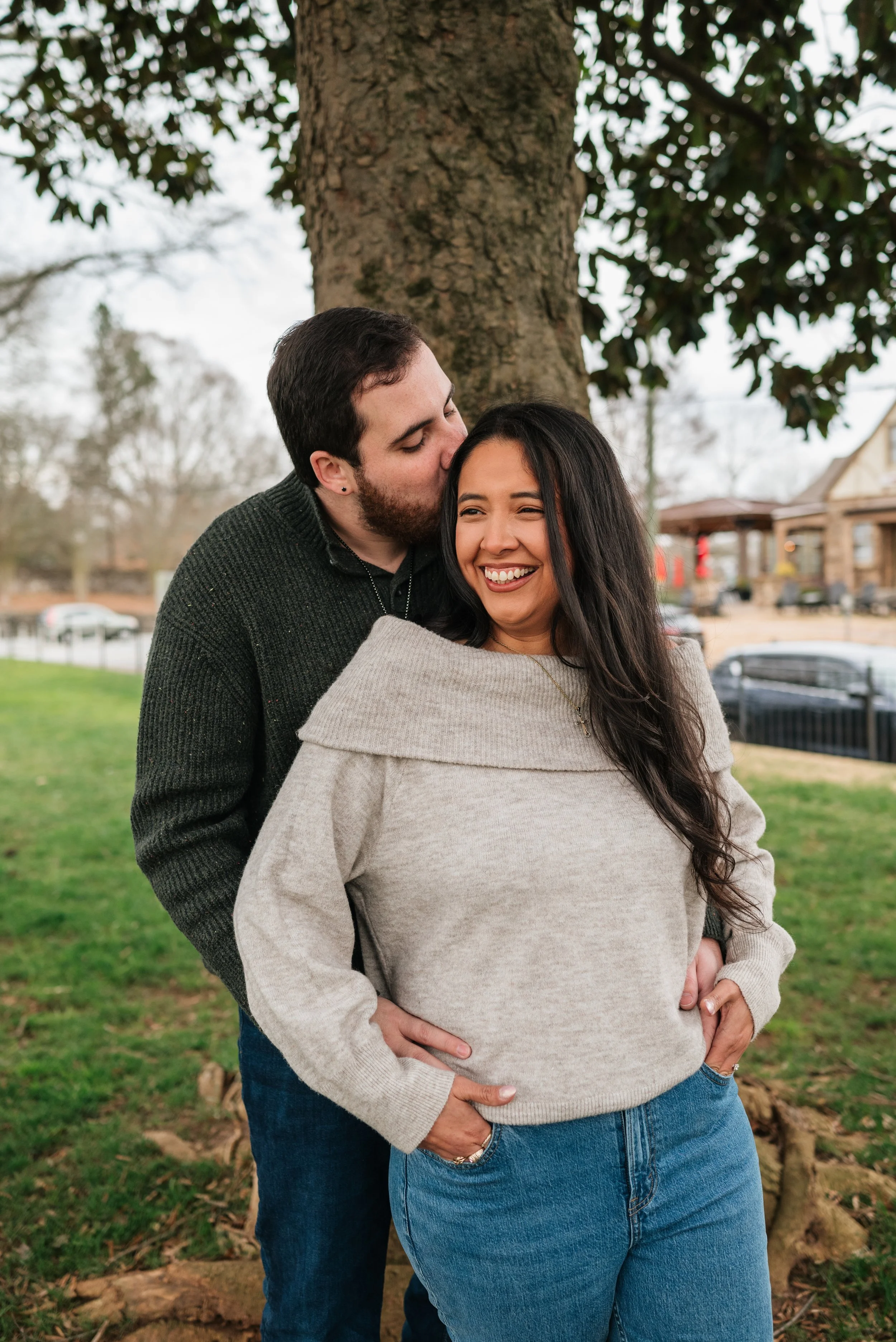 A man giving a kiss on the forehead to a woman smiling happily outdoors near a tree during their engagement photoshoot in Downtown Roswell, GA.