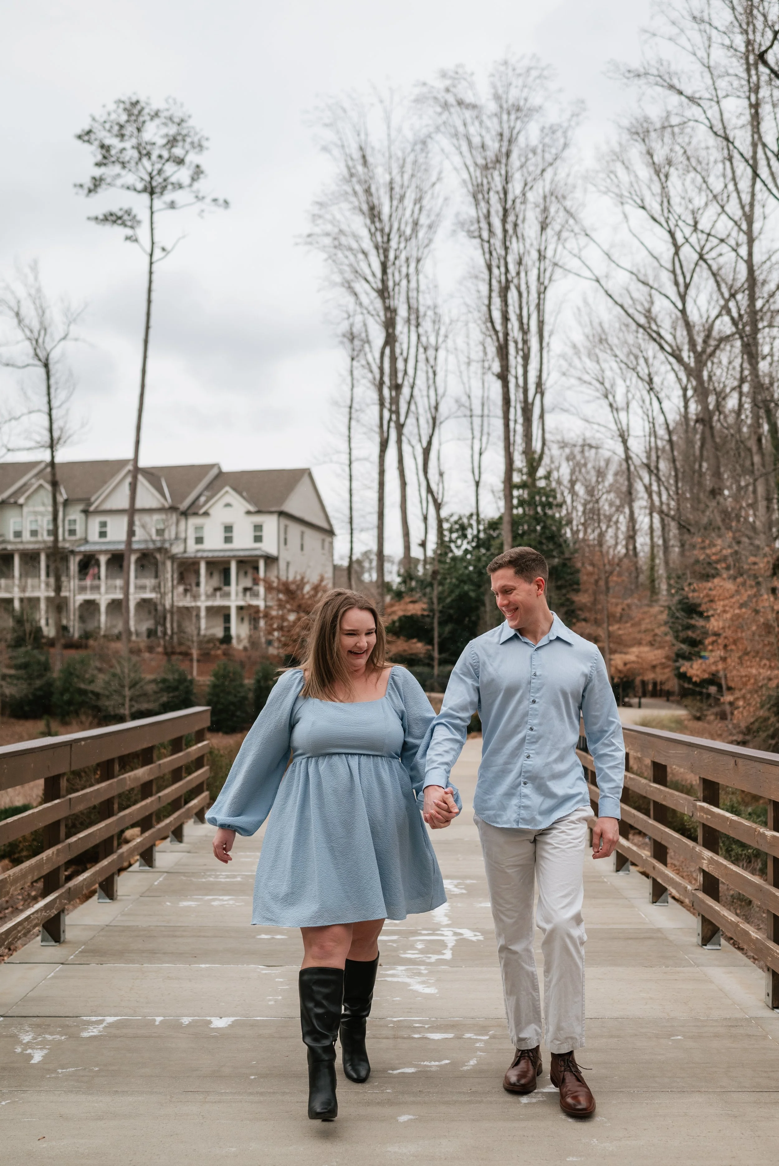 A couple holding hands and walking on a wooden bridge outdoors during late autumn or winter, with house and trees in the background during their engagement photoshoot in Alpharetta, GA.