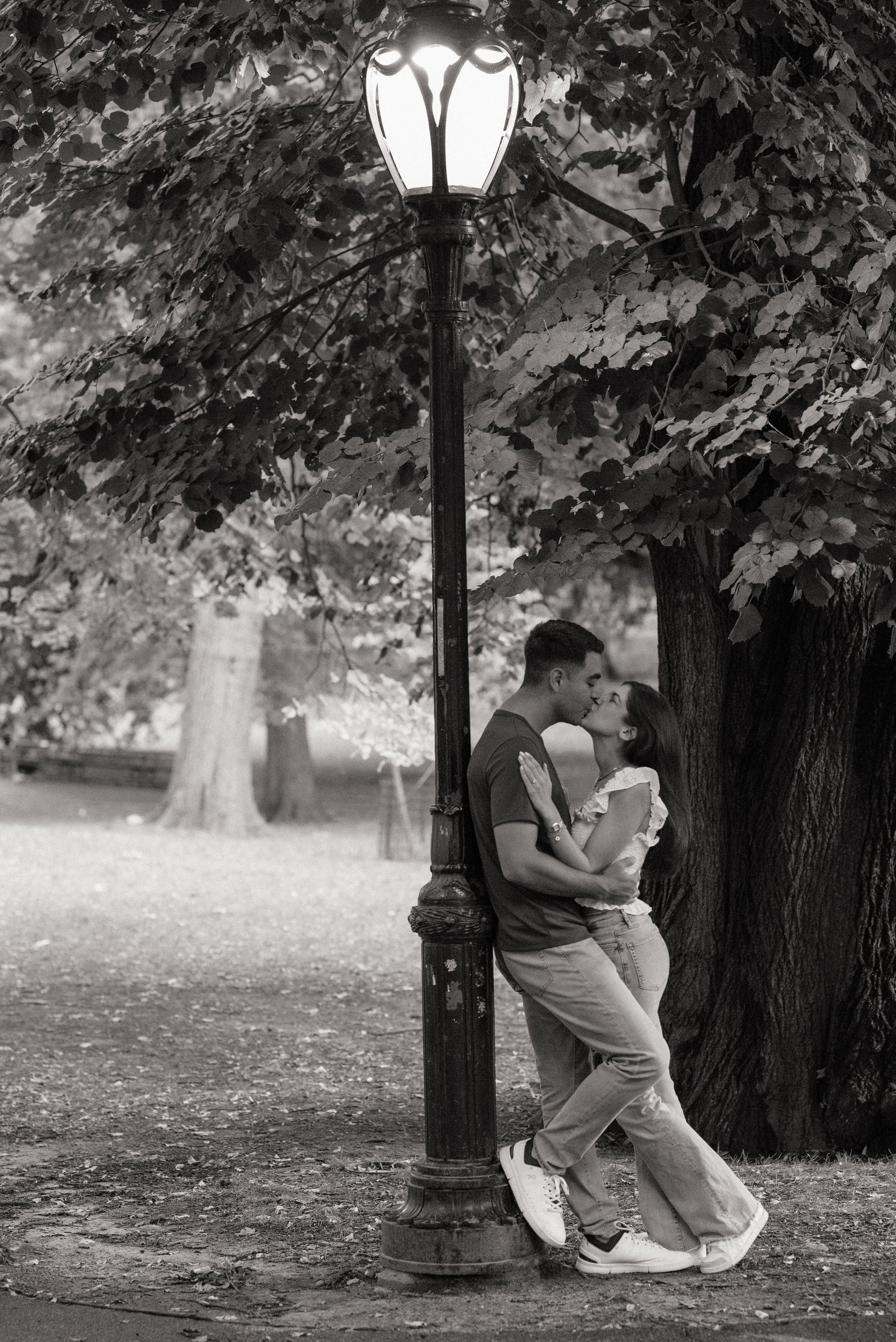 A black and white photo of a young couple kissing outdoors, leaning against a street lamp post under trees in a park.