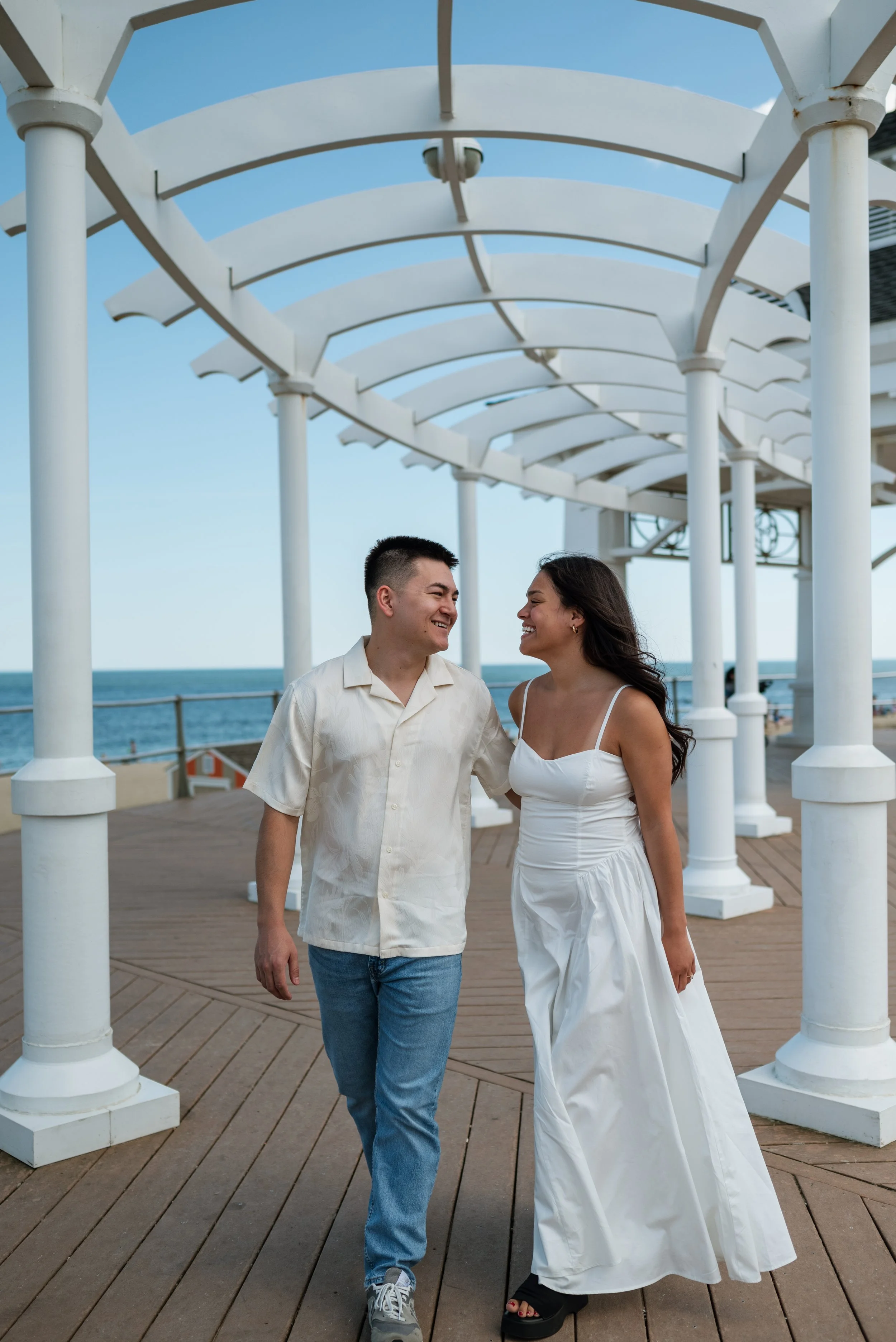 A happy couple walking together on a pier near the ocean, with a white pergola overhead and blue sky in the background.