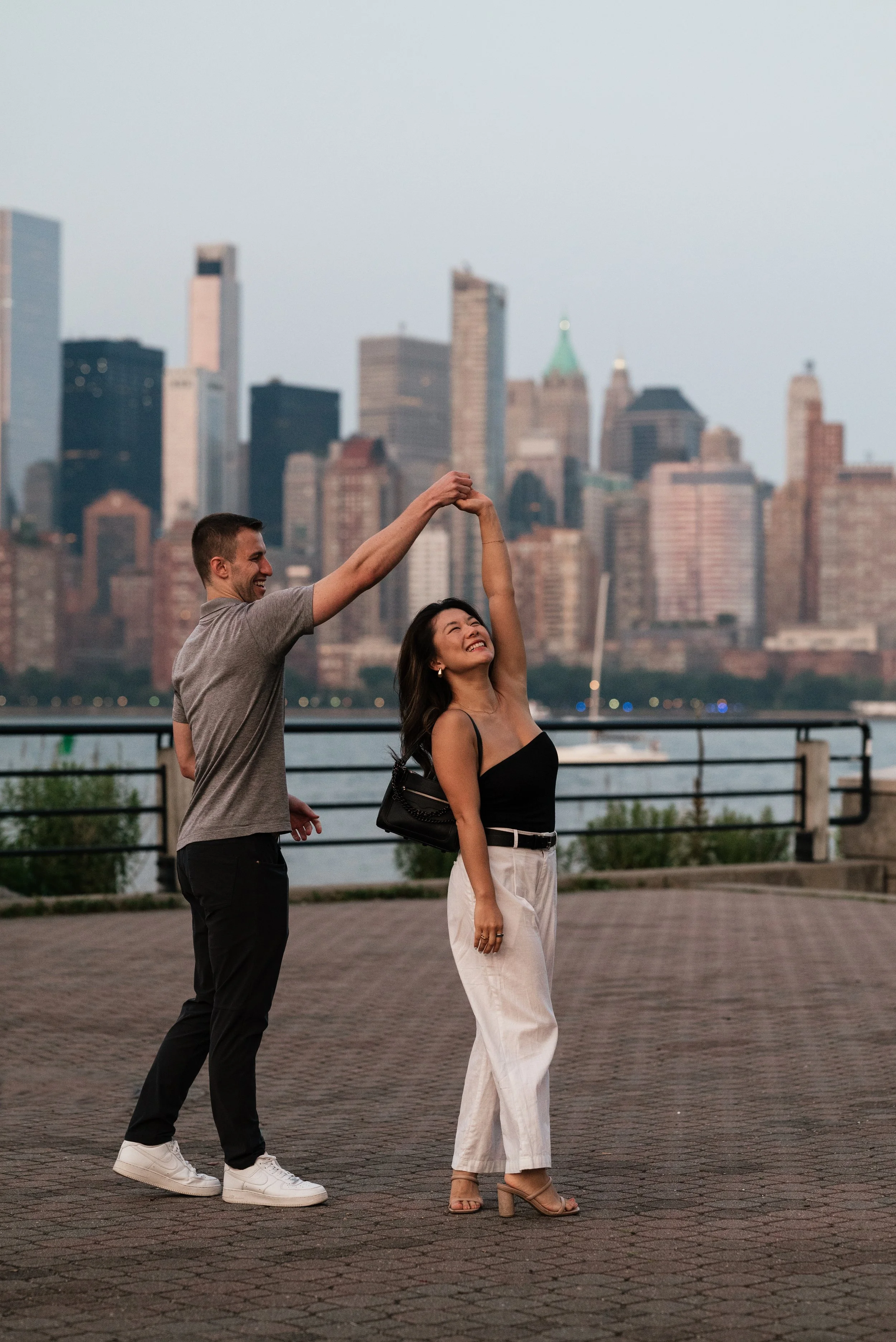 A man and woman dancing together outdoors near a river with a city skyline in the background, during evening or dusk.