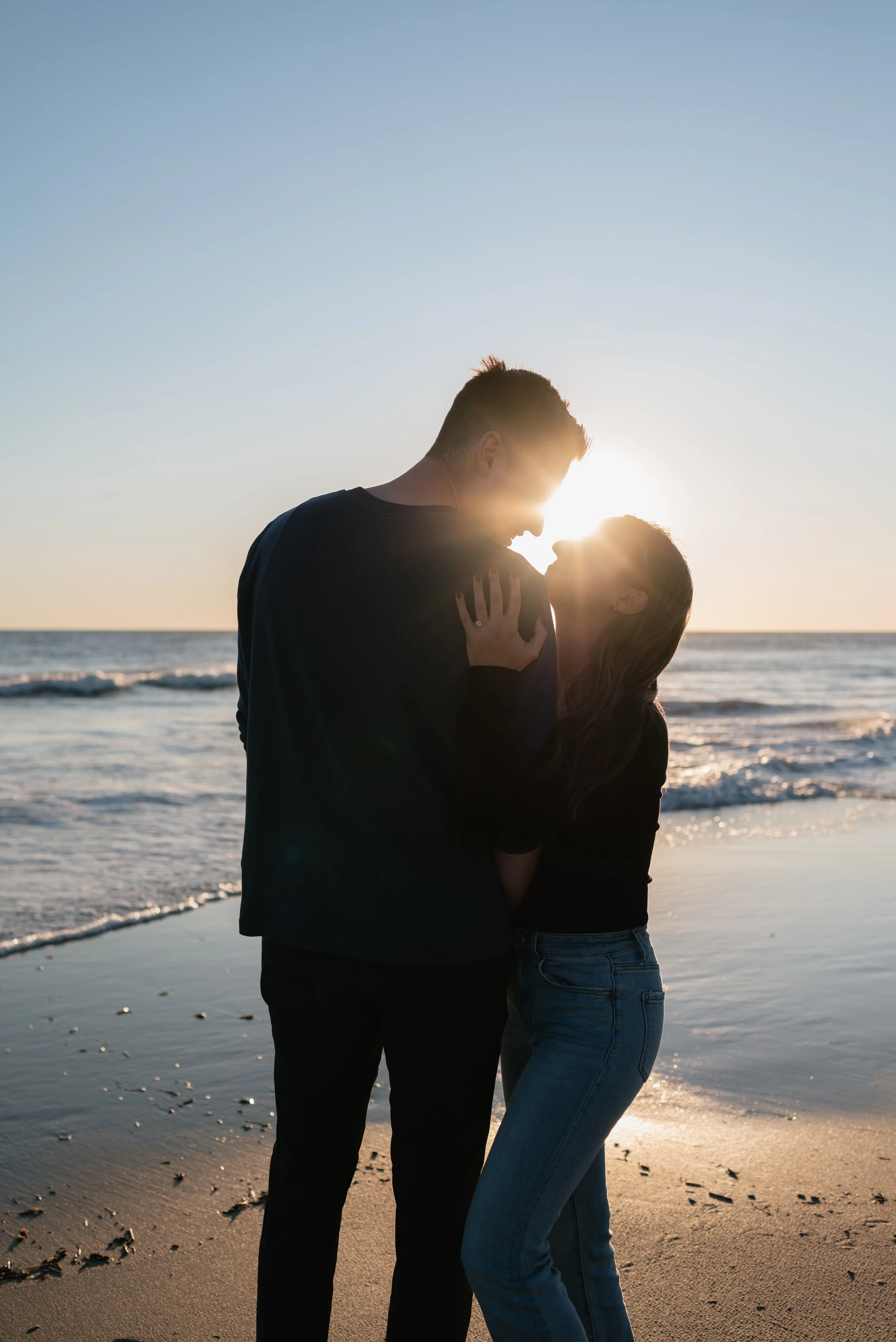 A couple embracing and sharing a kiss on the beach during sunset, with the sun setting over the ocean in the background.