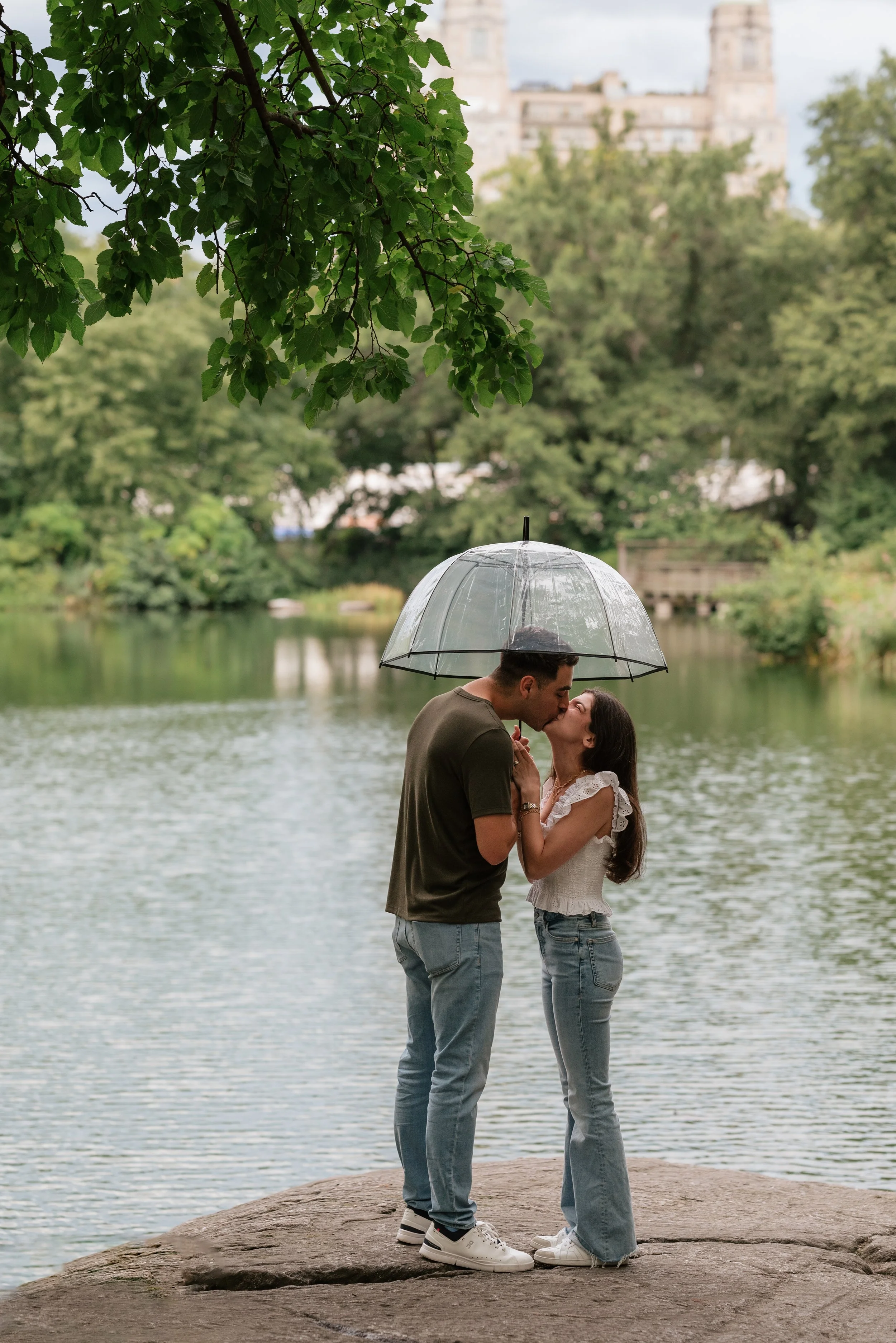 A couple sharing a kiss under a clear umbrella near a lake with trees in the background.