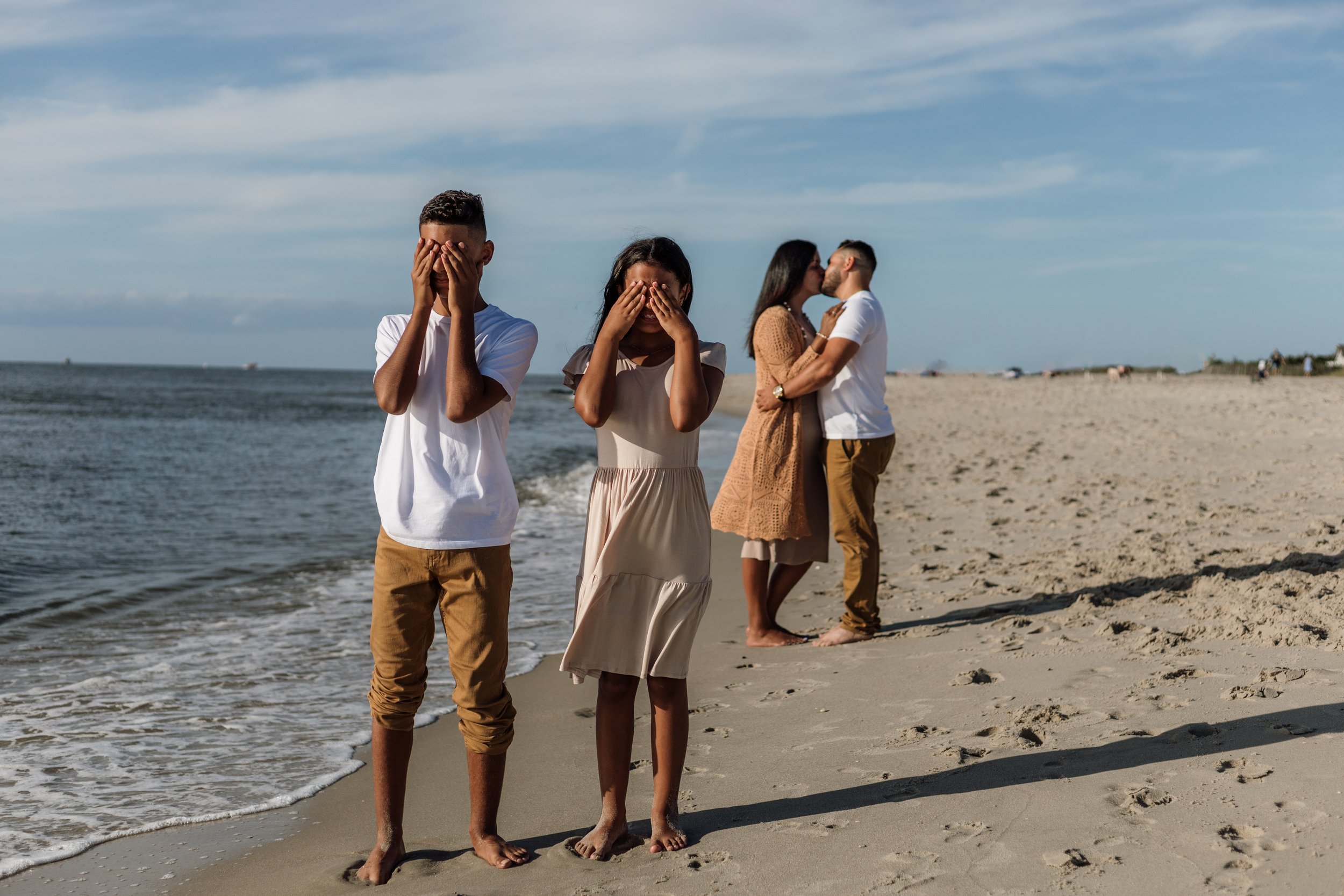 Two young women and two young men standing on a sandy beach, with the women covering their faces and the couples in the background kissing.