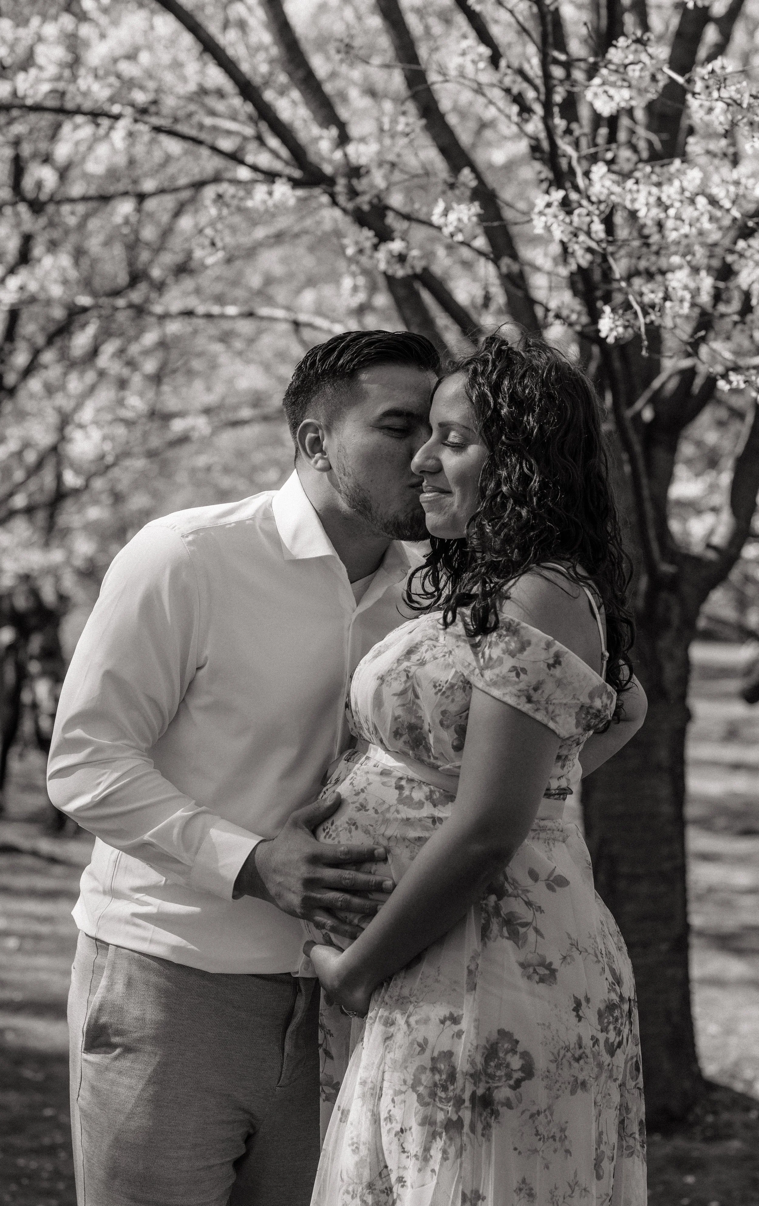 A black-and-white photo of a couples embracing outdoors under blooming trees; the man is kissing the woman on her forehead, and they are holding hands.