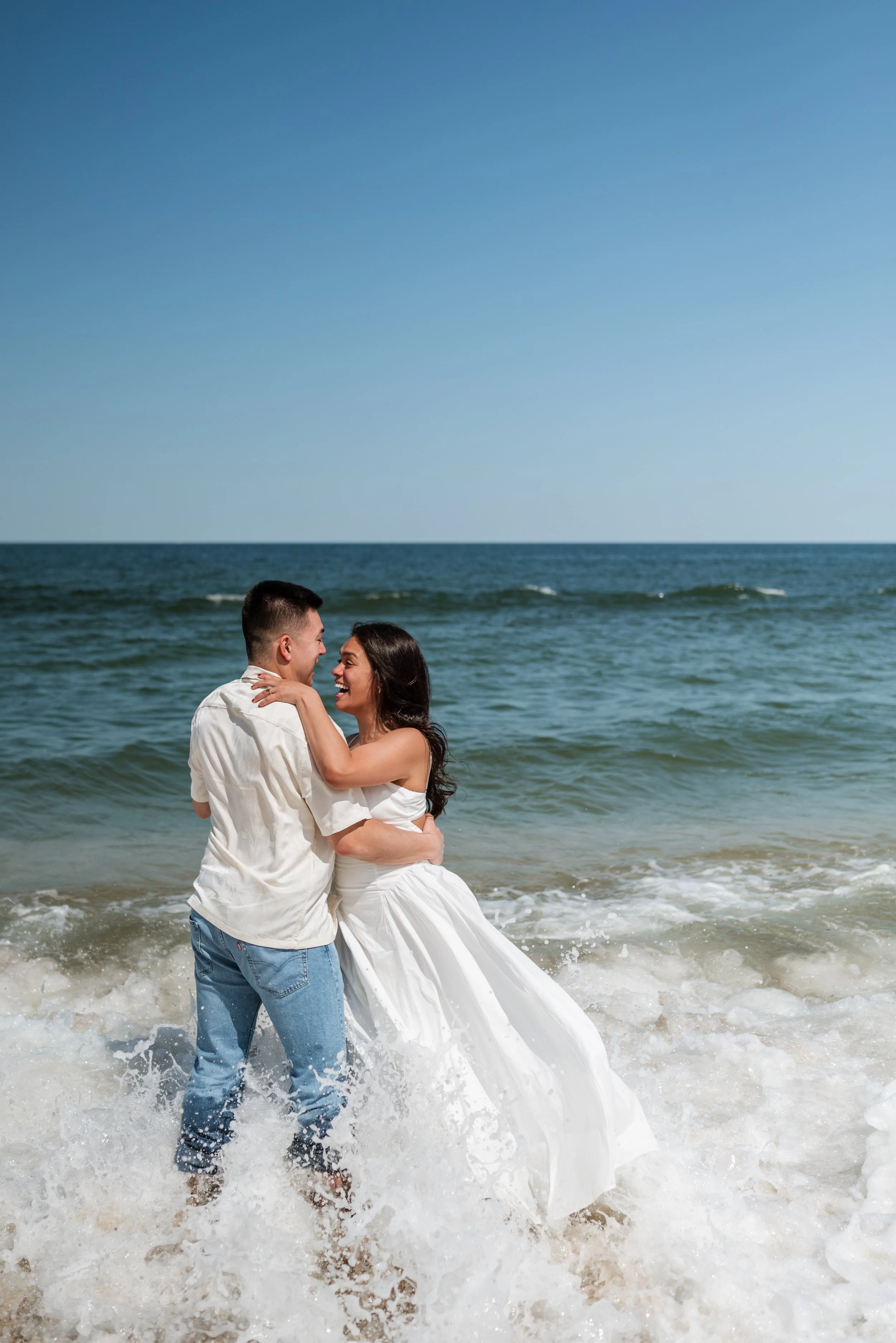 A couple dancing and laughing in the ocean waves on a sunny day, with a clear blue sky in the background.