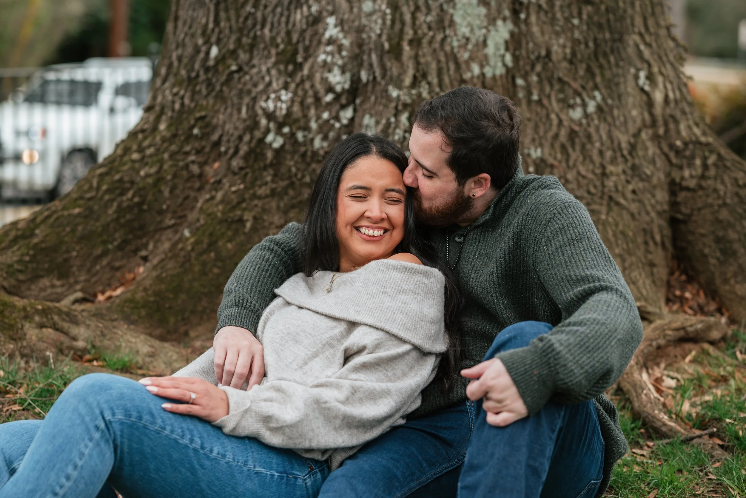 A couple sitting on grass in Downtown Roswell near a large tree, with the fiancé kissing his fiancé's forehead. The woman is smiling with her eyes closed, wearing a light gray sweater, and the man is wearing a dark green sweater.