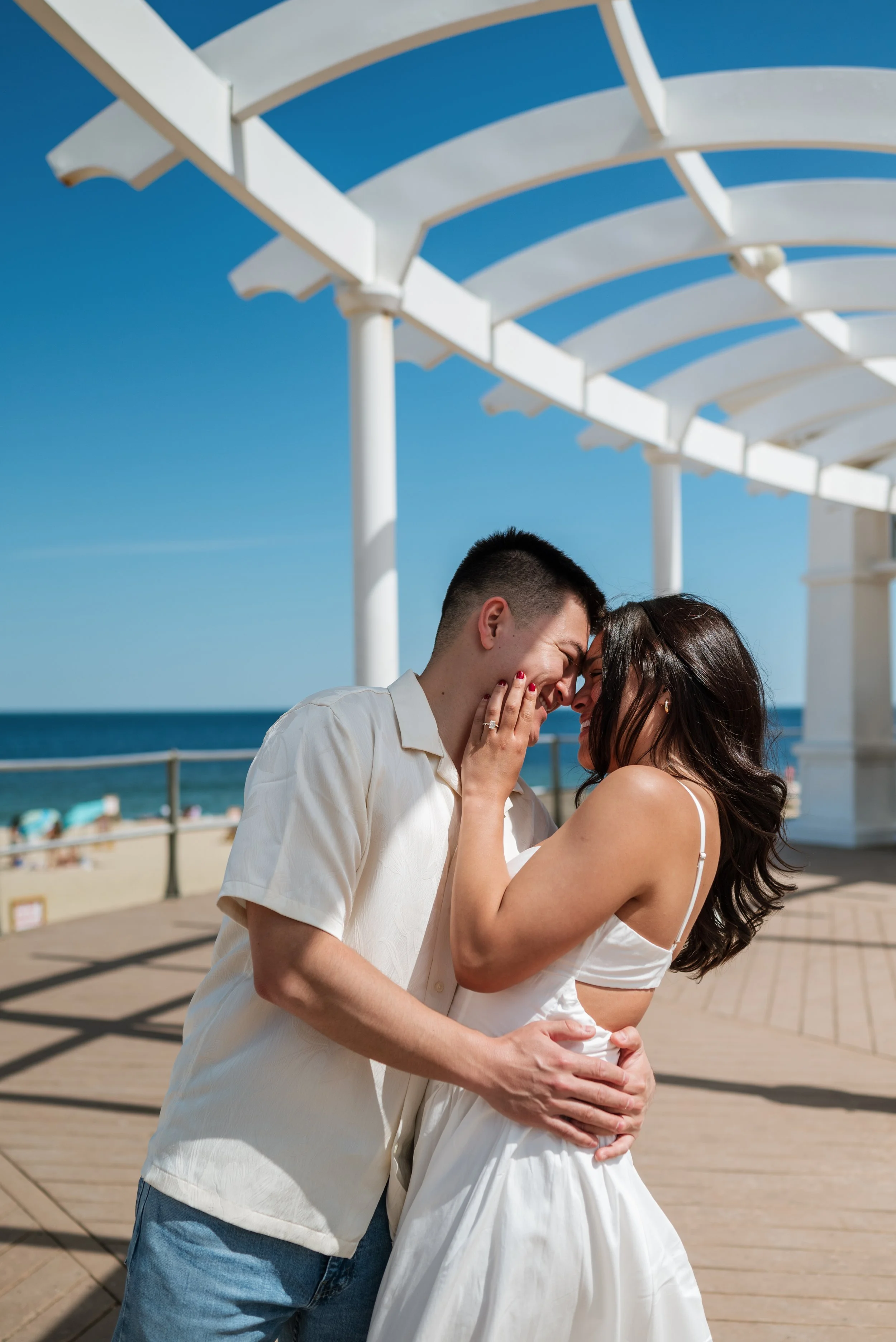 A happily smiling couple touching foreheads on a beachside boardwalk under a white pergola, with the ocean and sandy beach in the background, during a sunny day.