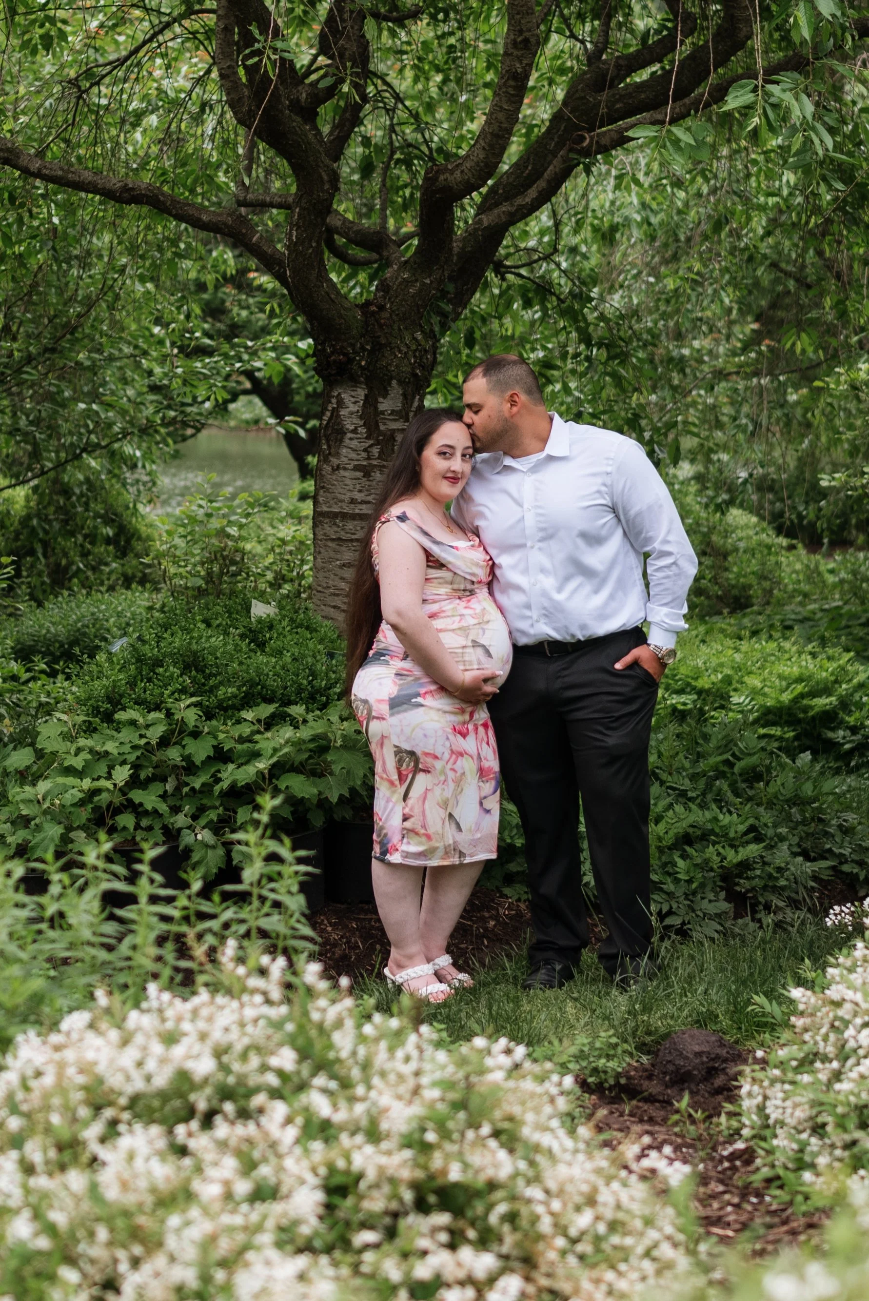 A pregnant woman and a man are standing together outdoors near a tree and greenery, with the man giving her a kiss on the forehead.
