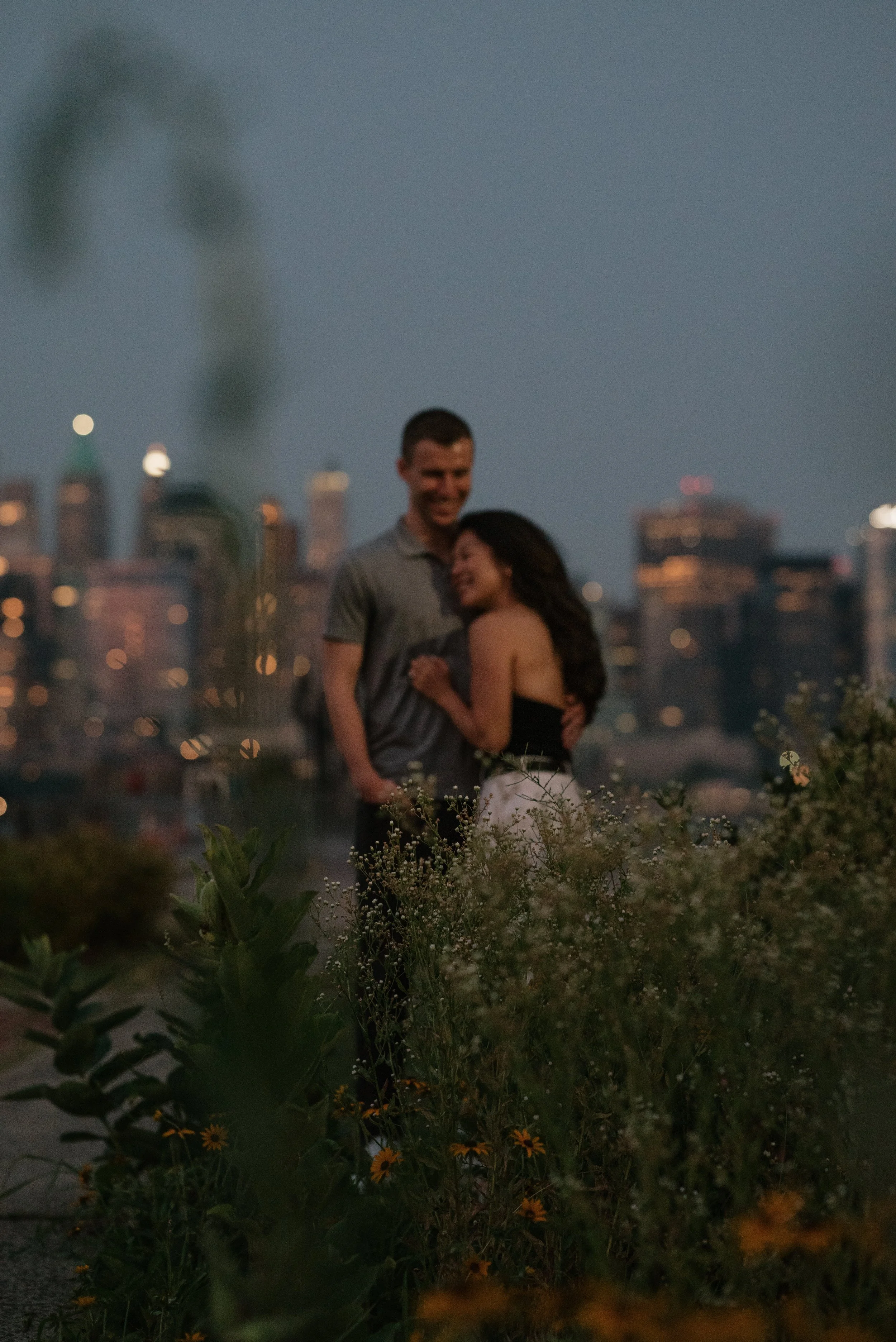 A couple standing together in a garden at dusk, with city buildings illuminated in the background.