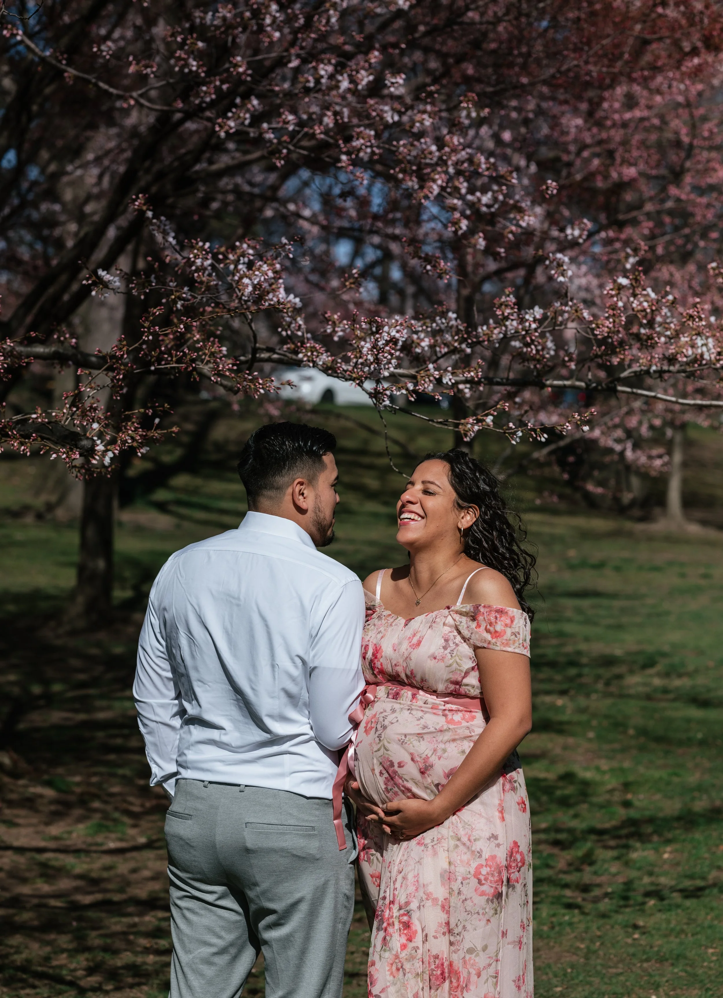 A pregnant woman and her partner smiling at each other under pink cherry blossom trees in a park.