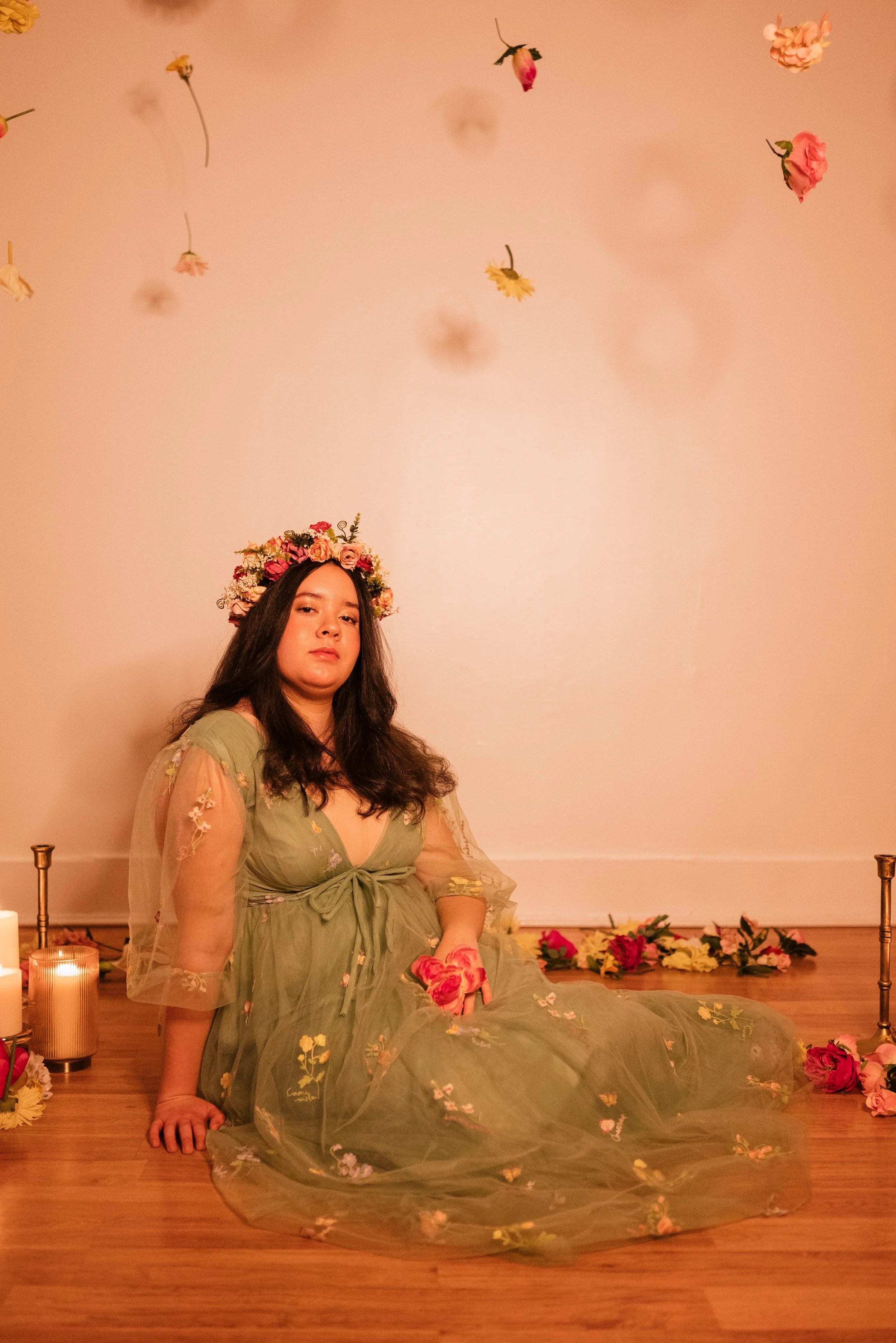 A woman in a green, floral dress with a transparent, embroidered overlay is sitting on the wooden floor with a subdued expression. She has long dark hair, a floral crown on her head, and holds a pink flower. The scene is decorated with hanging flower