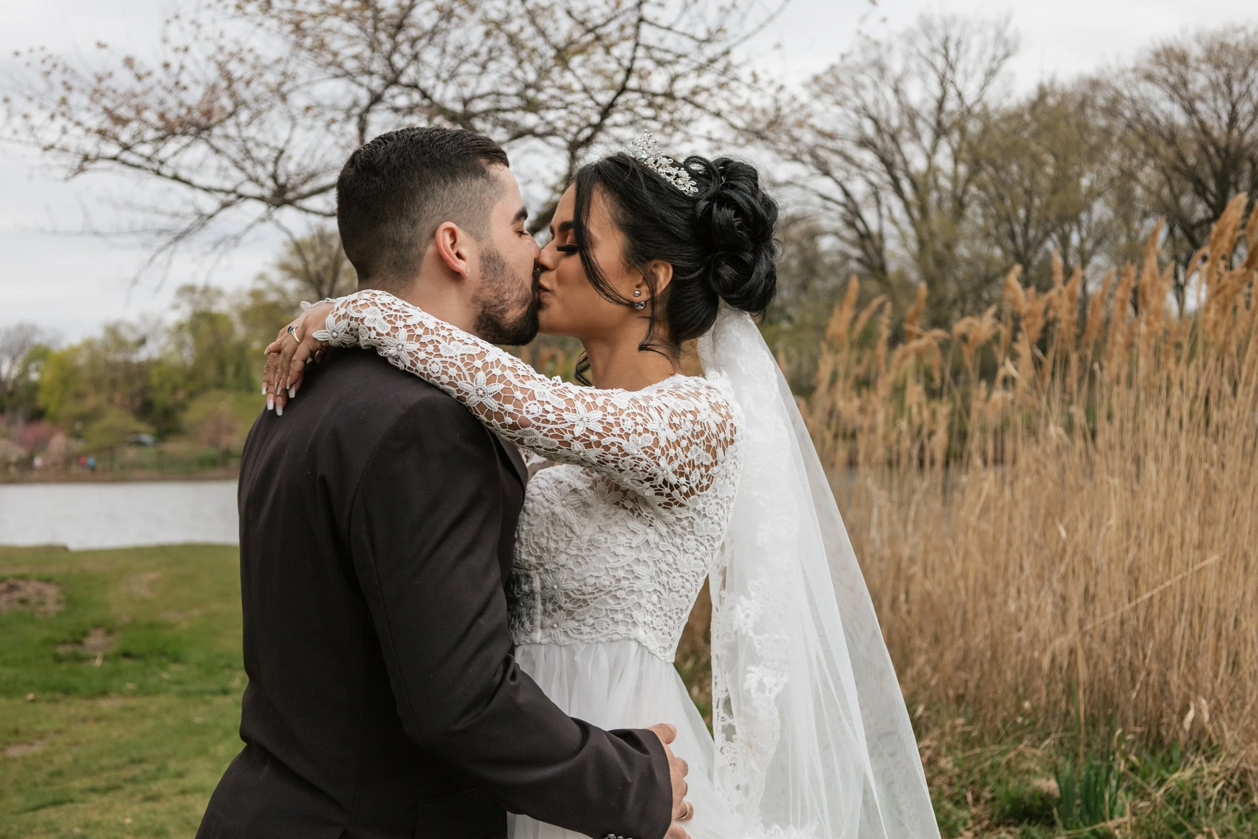 A newlywed couple sharing a kiss outdoors near a lake, with tall reeds and trees in the background.
