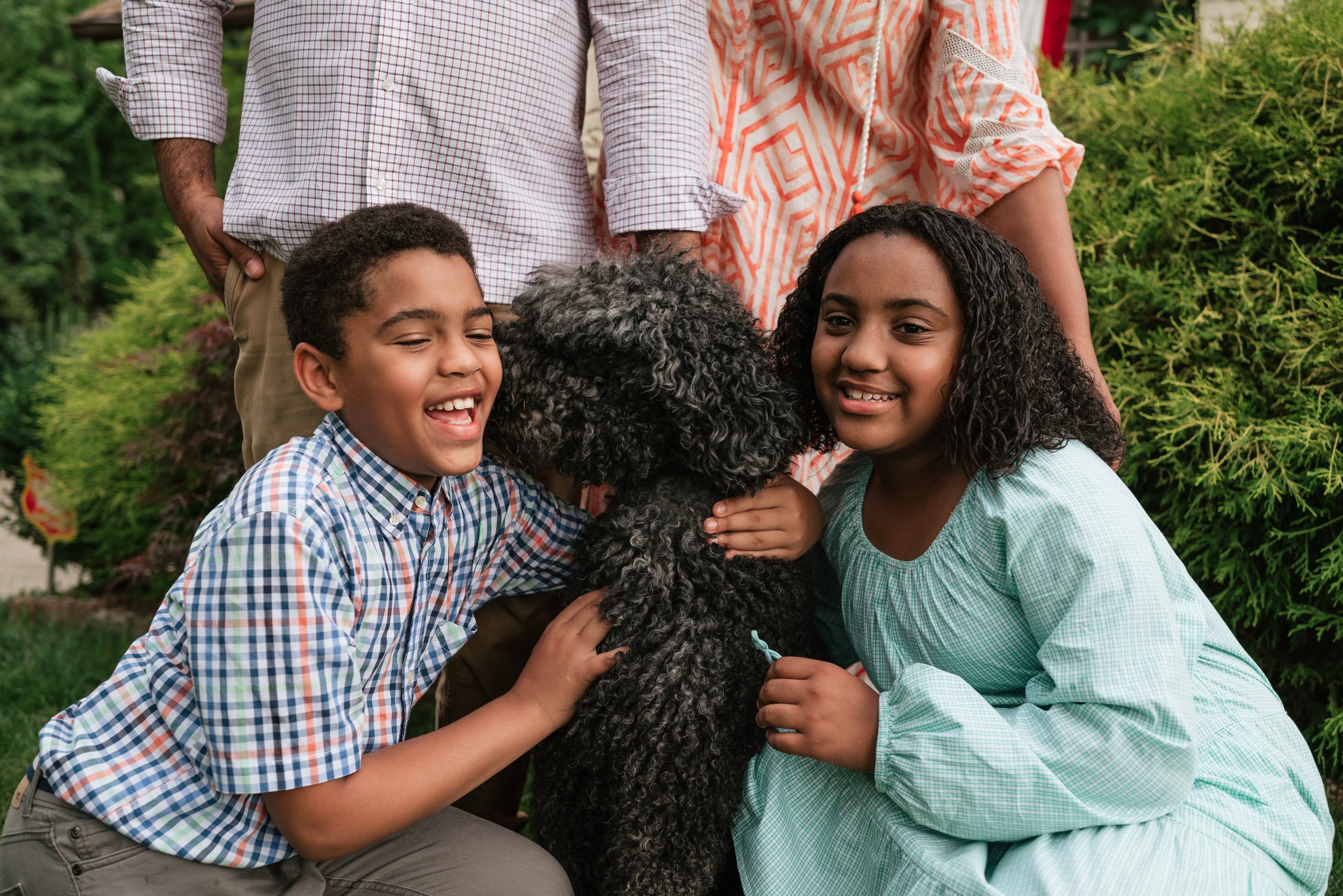 A group of children and two adults outside, smiling and hugging a black curly-haired dog.