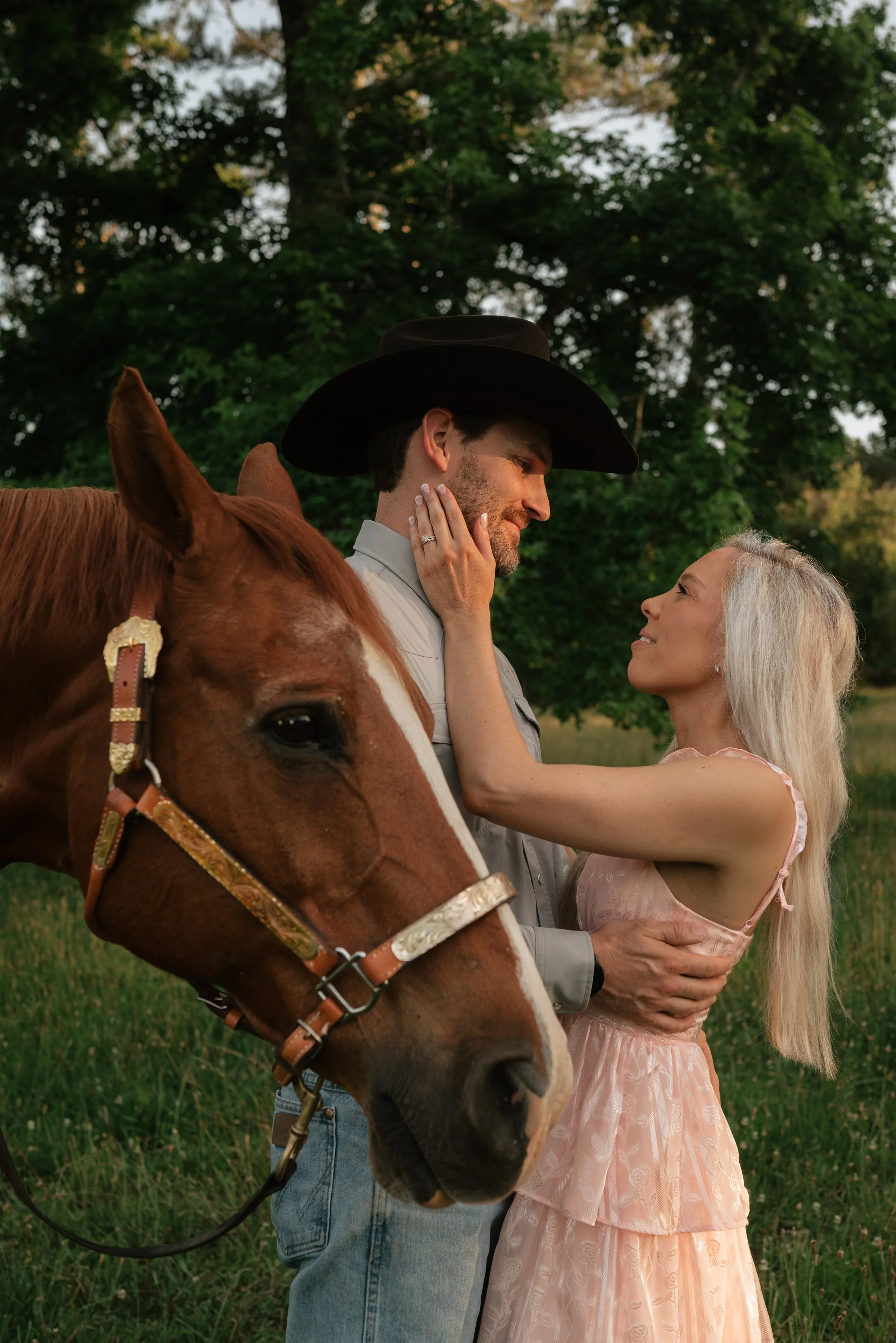 A man and woman standing close in a green outdoor setting, with the man wearing a black cowboy hat and the woman with long blond hair, gazing at each other. The man has a beard and wears a light-colored shirt, while the woman wears a pink dress. A ho