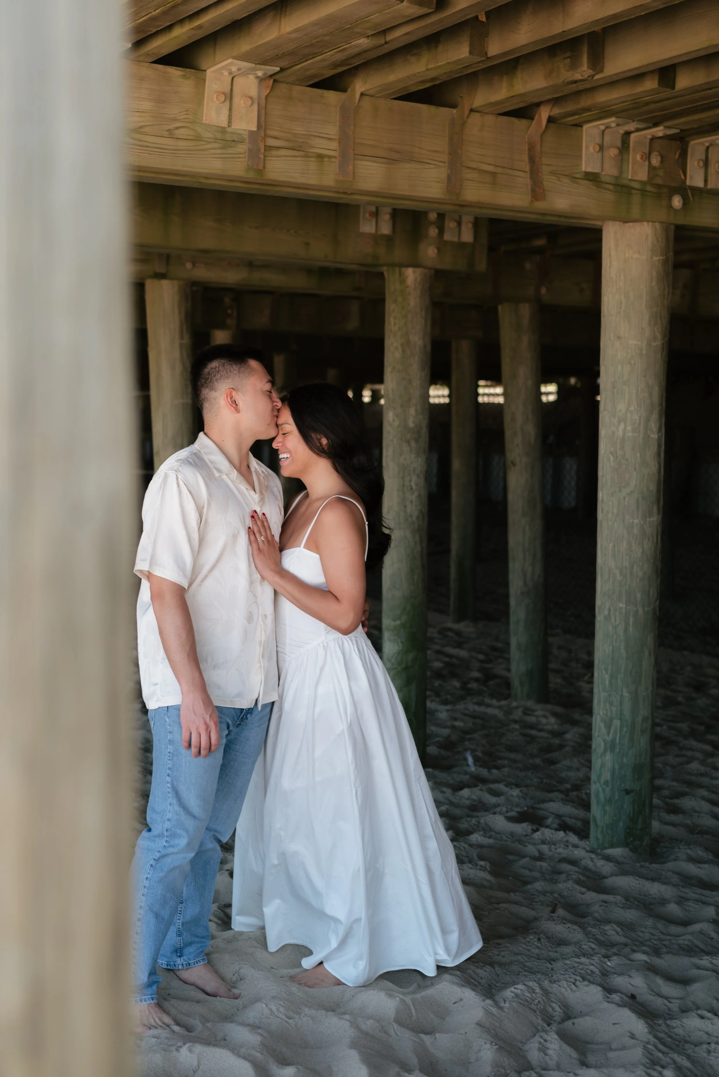 A couple standing on the beach underneath a wooden pier, sharing an intimate moment with the man kissing the woman's forehead and the woman smiling happily.