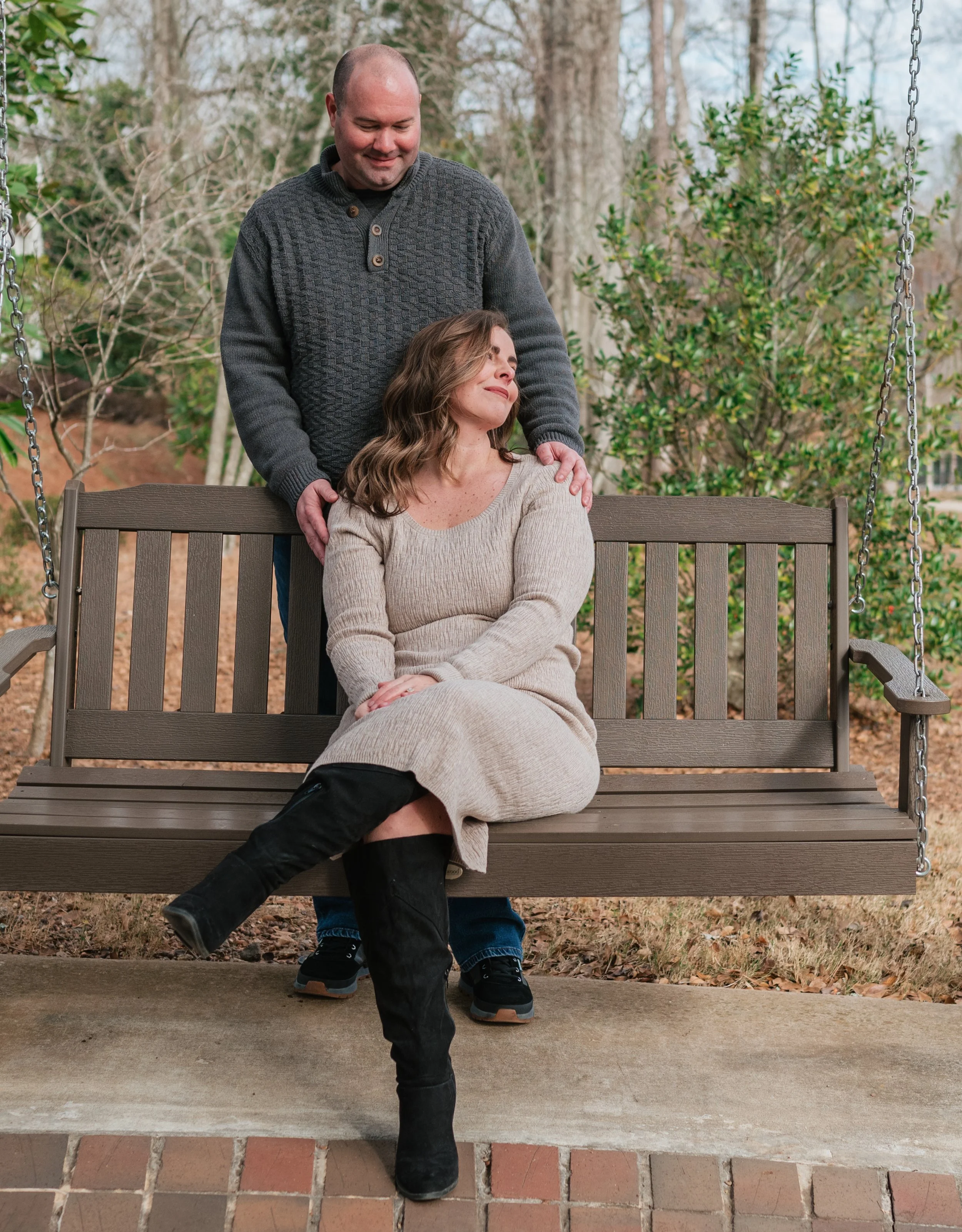 A man standing behind a seated woman on a porch swing, with one hand on her shoulder and the other on her back, smiling as she looks to the side. The woman is wearing a beige dress and black knee-high boots, while the man is wearing a dark sweater an