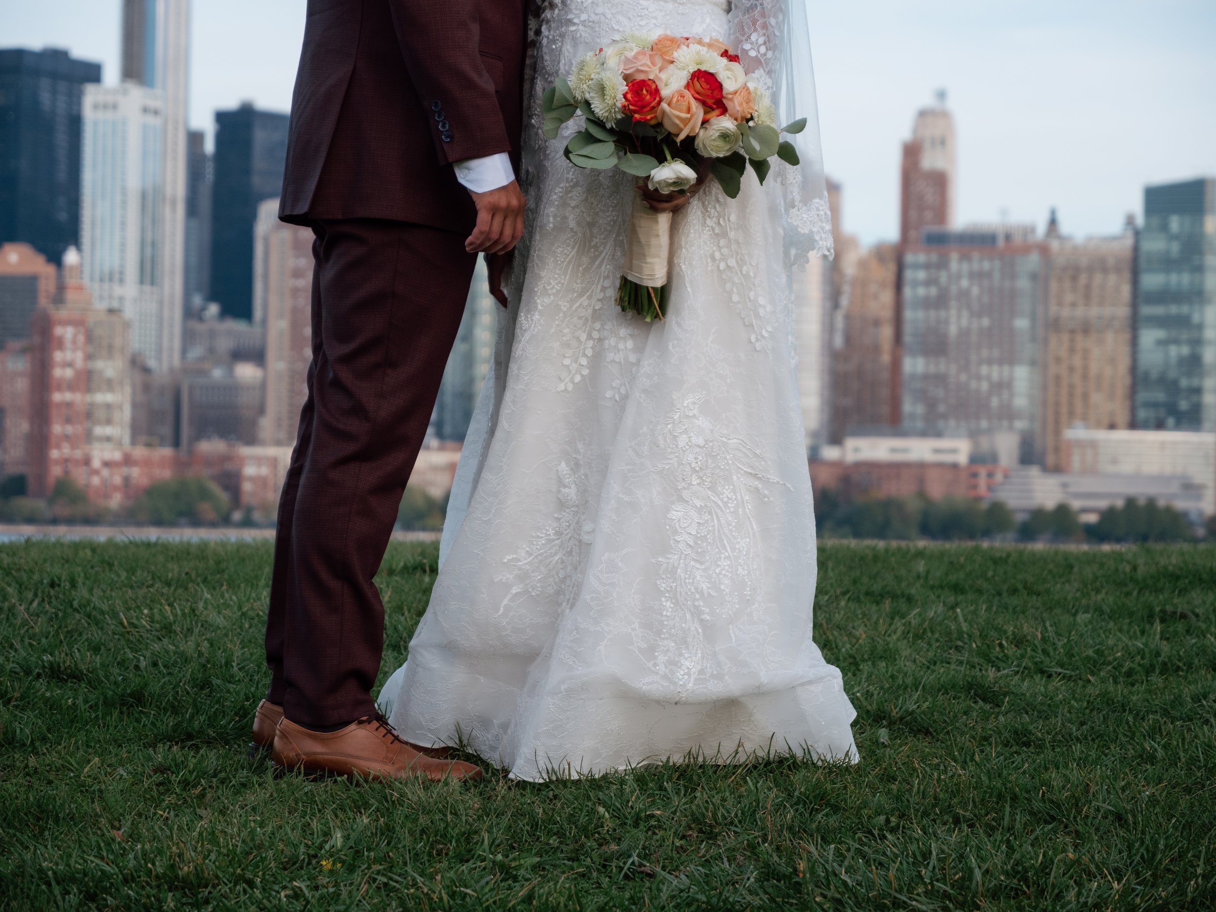 Bride and groom standing on grass with city skyline in background, the bride holding a bouquet of flowers.