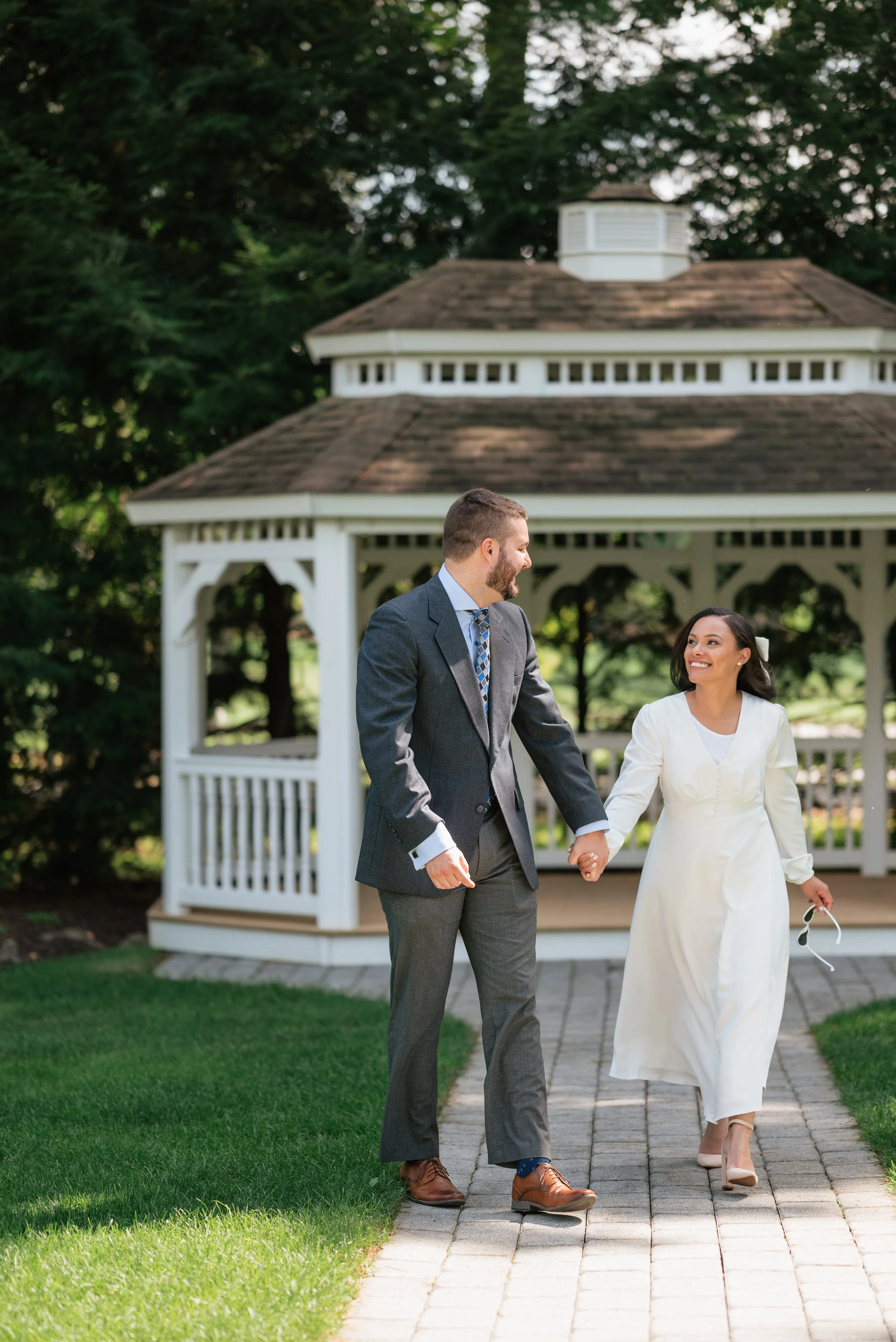 A couple is walking hand in hand outdoors near a white gazebo surrounded by greenery. The man is wearing a gray suit and the woman is dressed in a white dress, smiling at each other.
