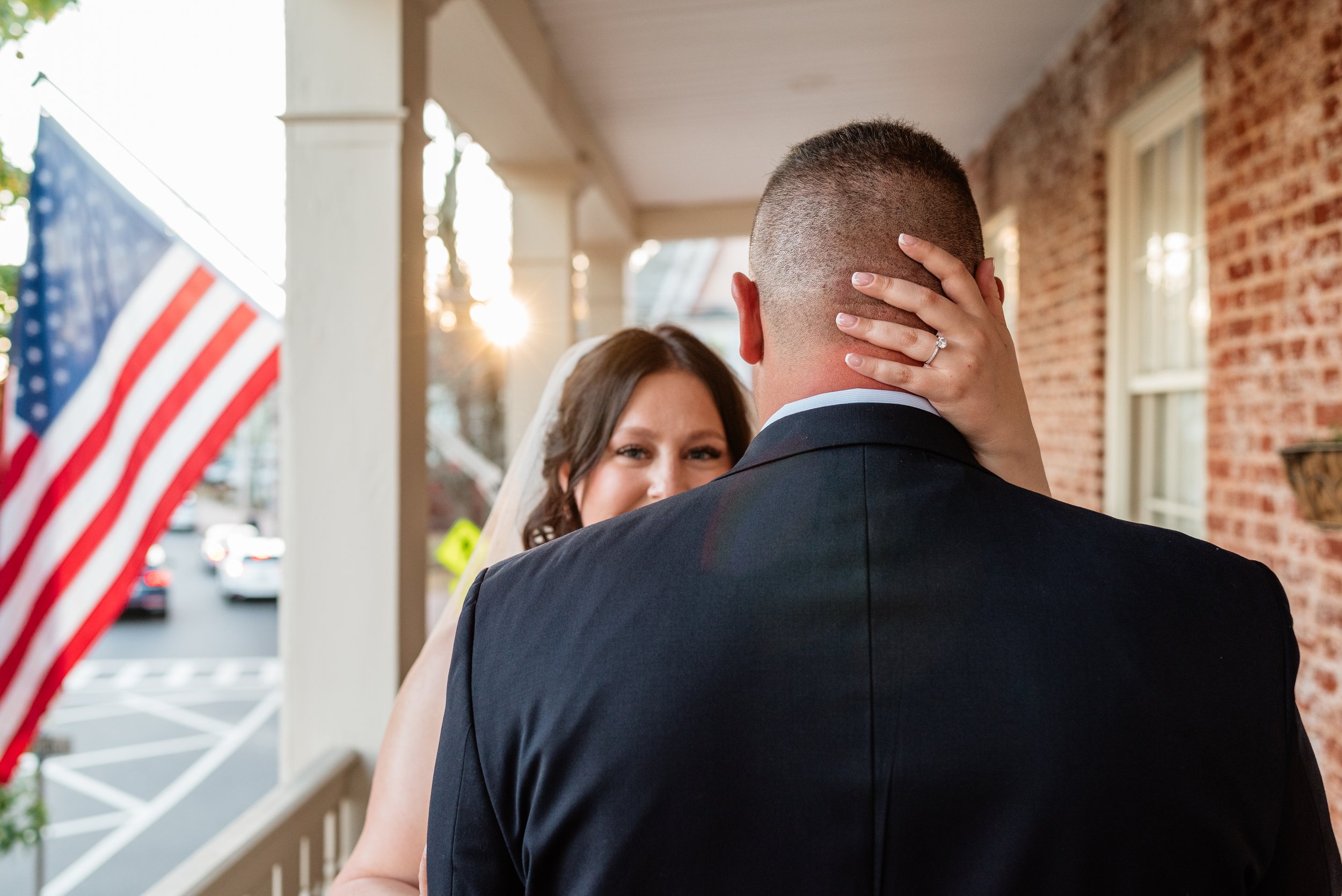 A bride and groom share a kiss on a porch, with the bride's hand on the back of the groom's head, showing her wedding ring, as the bride looks into his eyes.