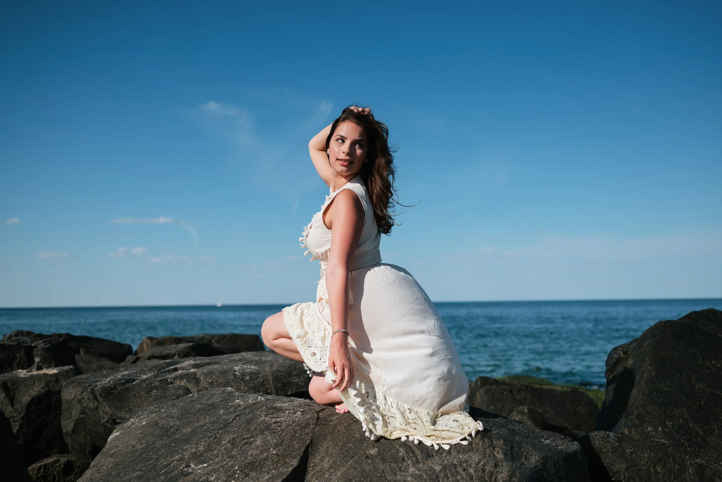 A woman in a white dress kneeling on rocks by the ocean under a clear blue sky.