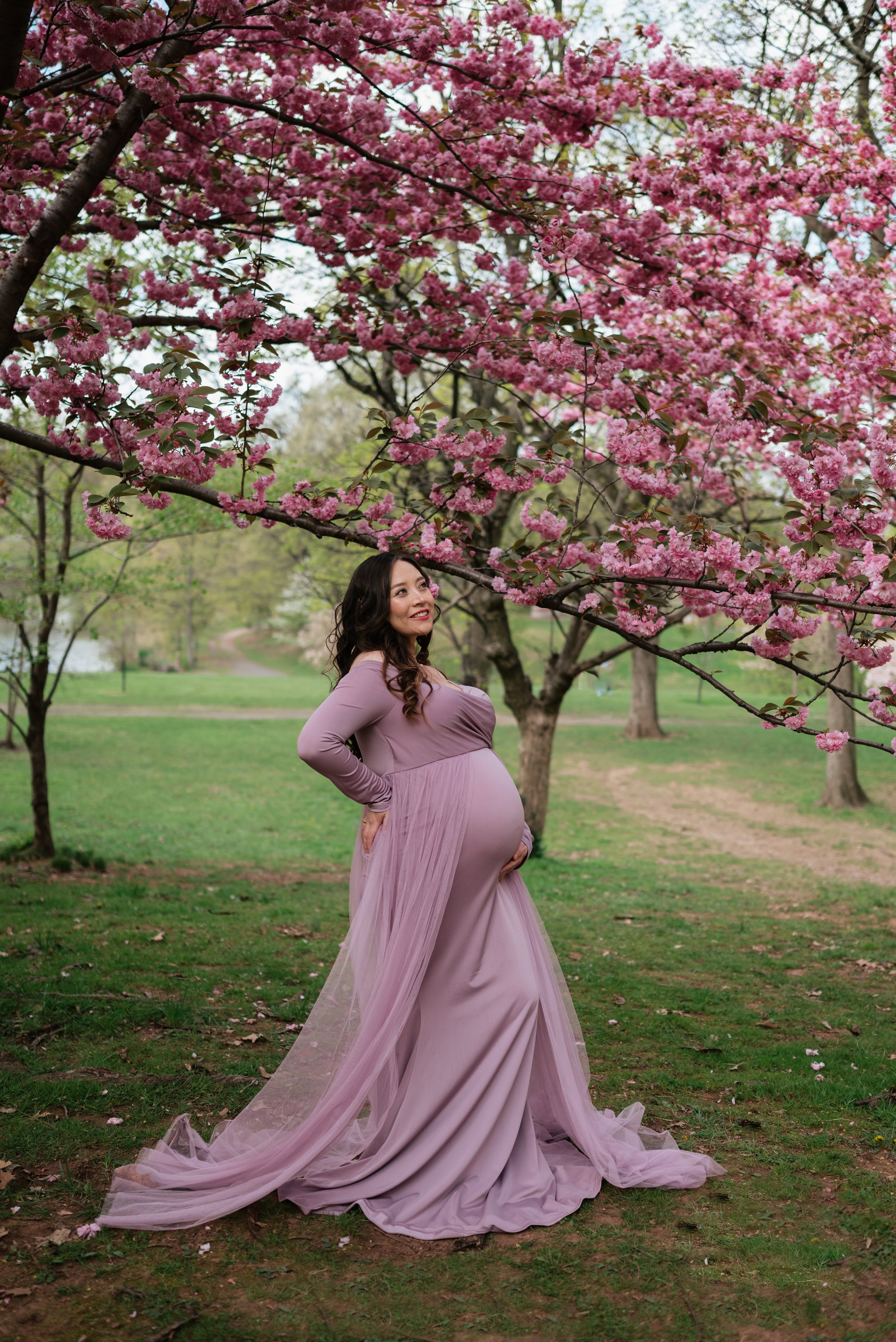 A pregnant woman in a mauve dress poses outdoors under pink cherry blossom trees, smiling and gently touching her belly.