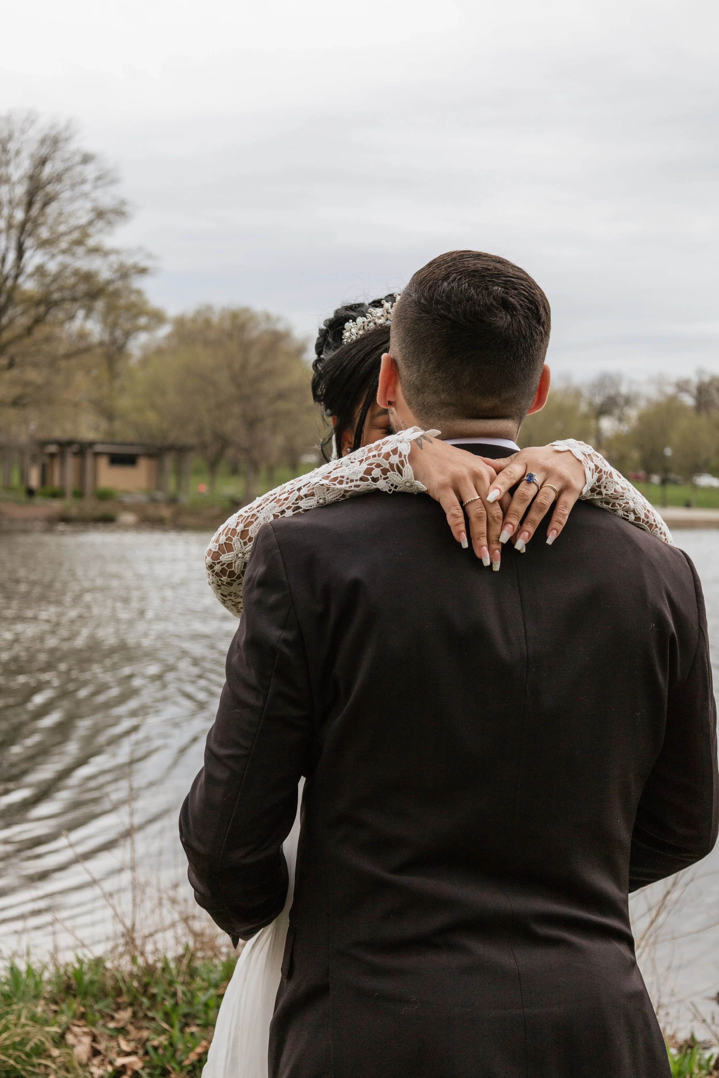 A couple dressed in wedding attire embracing by a lake, with trees and a cloudy sky in the background.
