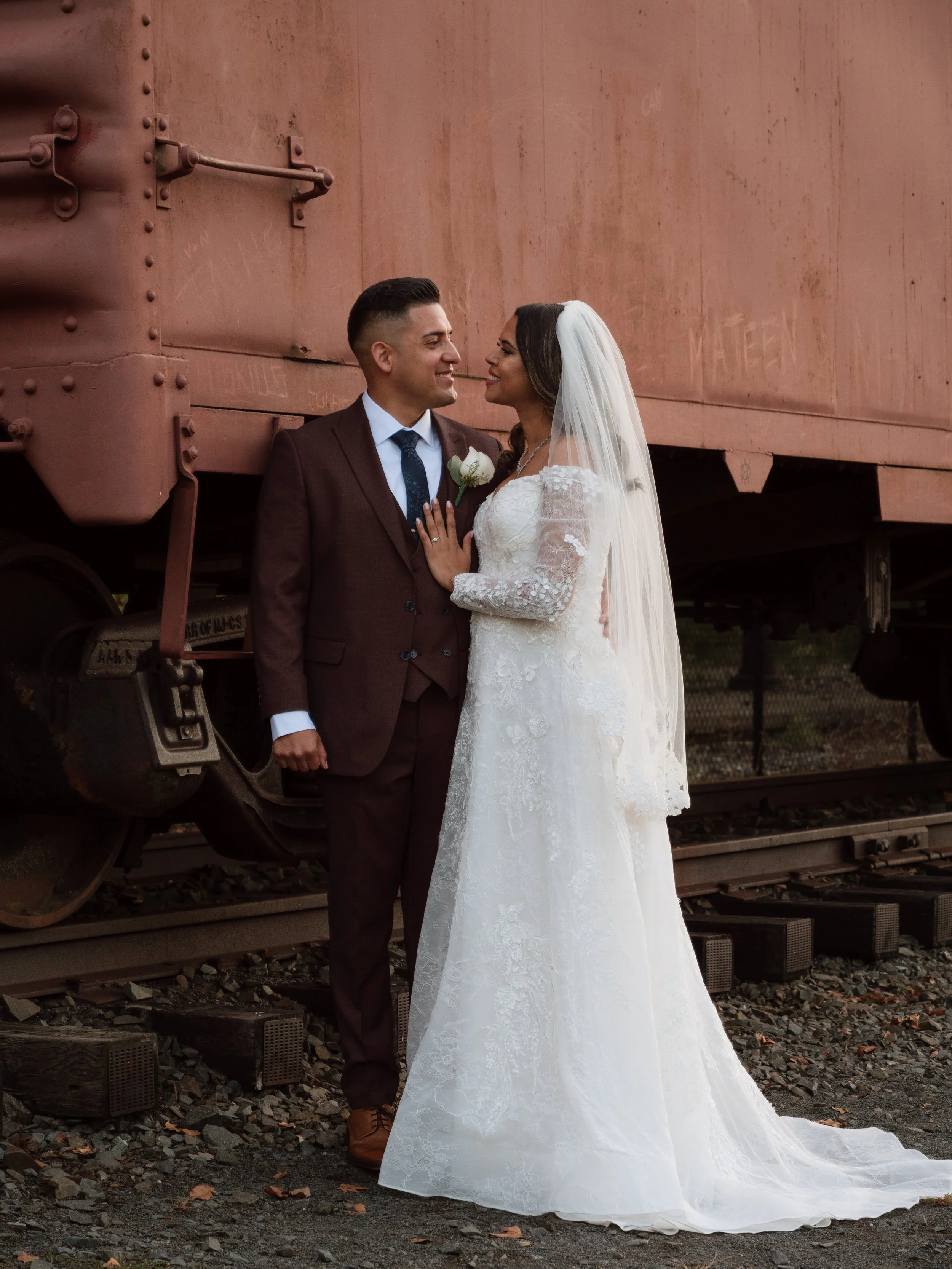 A bride and groom in wedding attire standing close together outdoors near a rusted train car, gazing into each other's eyes.