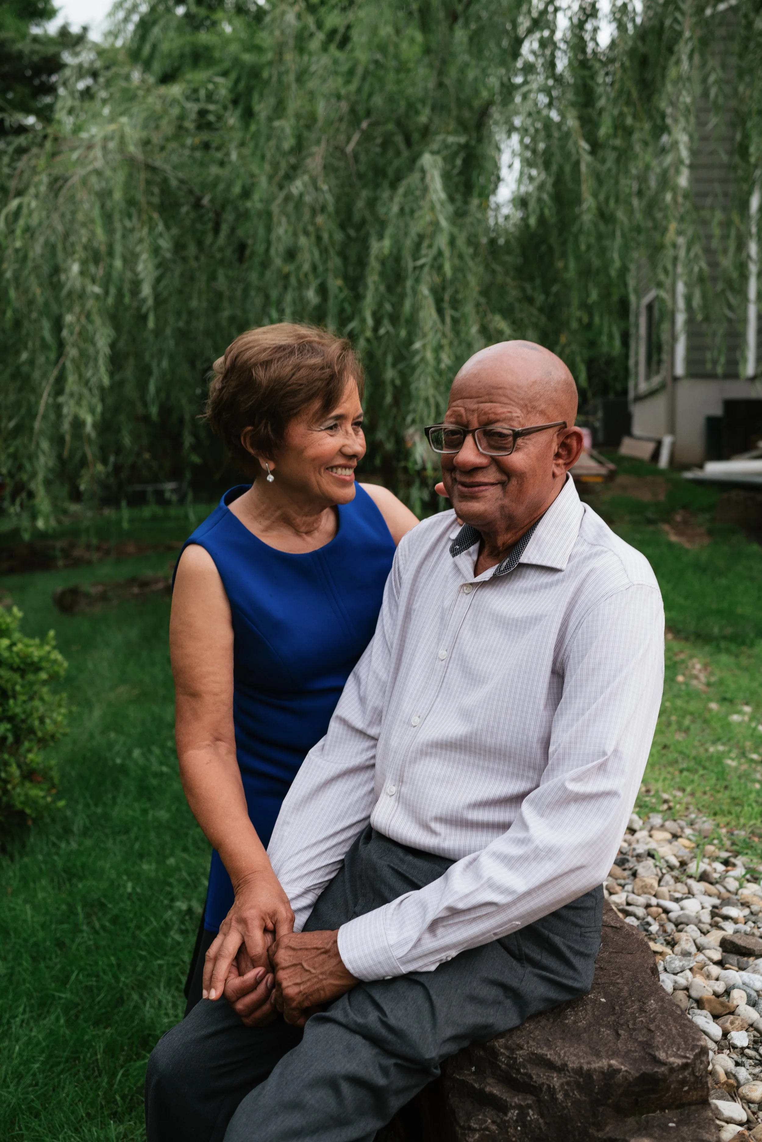 An elderly woman in a royal blue sleeveless dress smiling at an elderly man in a white shirt and dark pants, sitting outdoors on a rock with greenery and a house in the background.