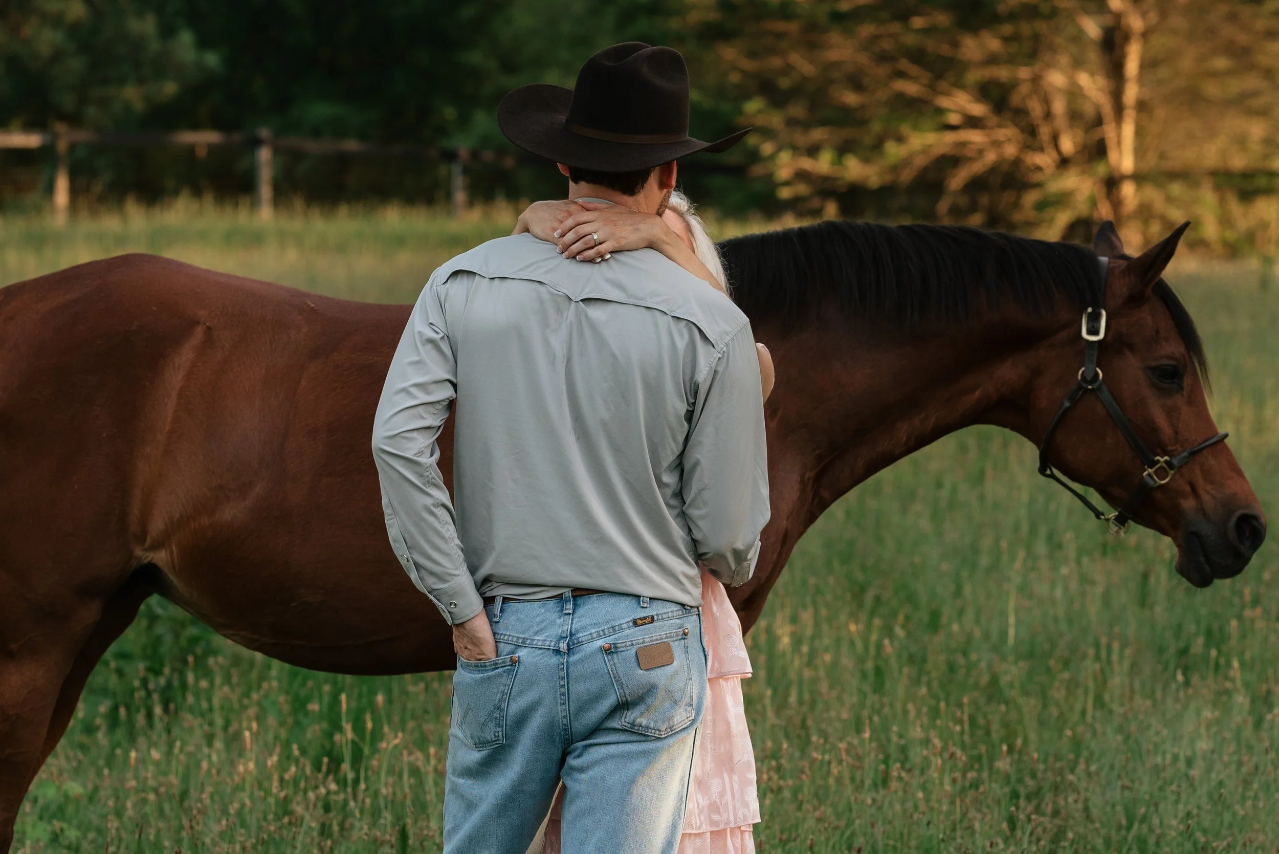 A couple embraces near a brown horse in a grassy field with trees in the background during sunset.