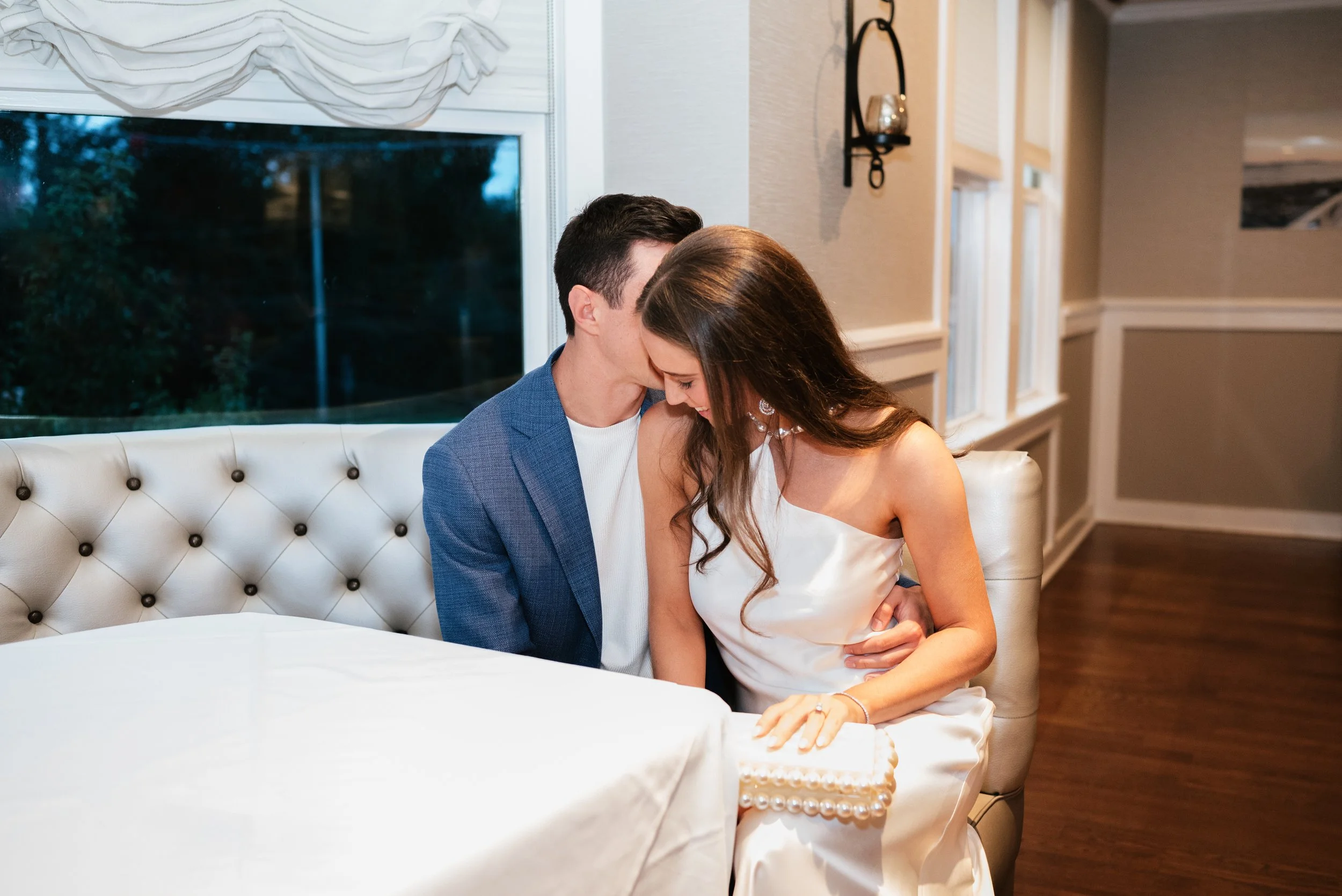 A couple sitting closely together on a white, tufted sofa, with their foreheads touching.