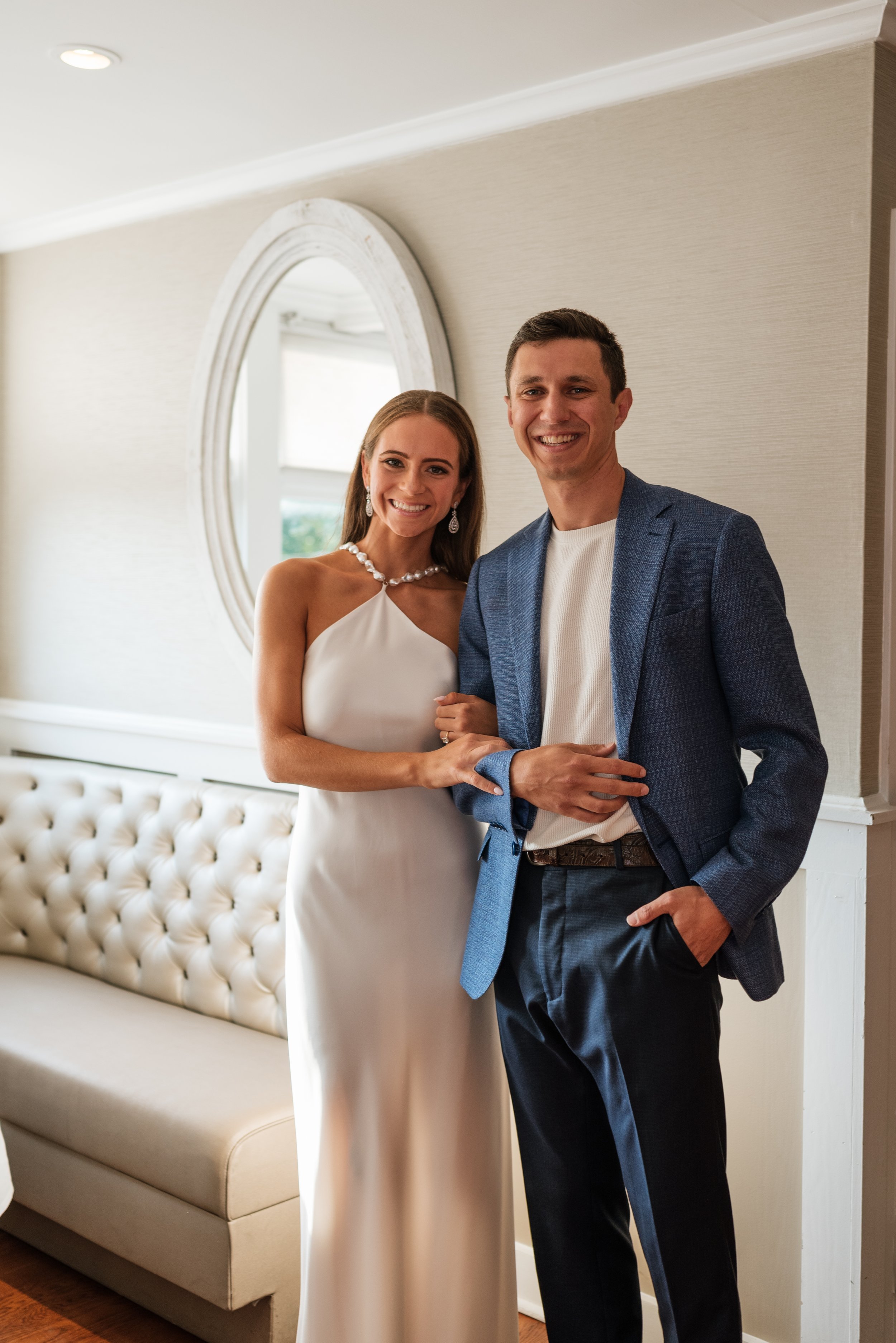 A woman in a white dress and pearl jewelry standing next to a man in a blue blazer and white shirt, holding hands and smiling indoors.