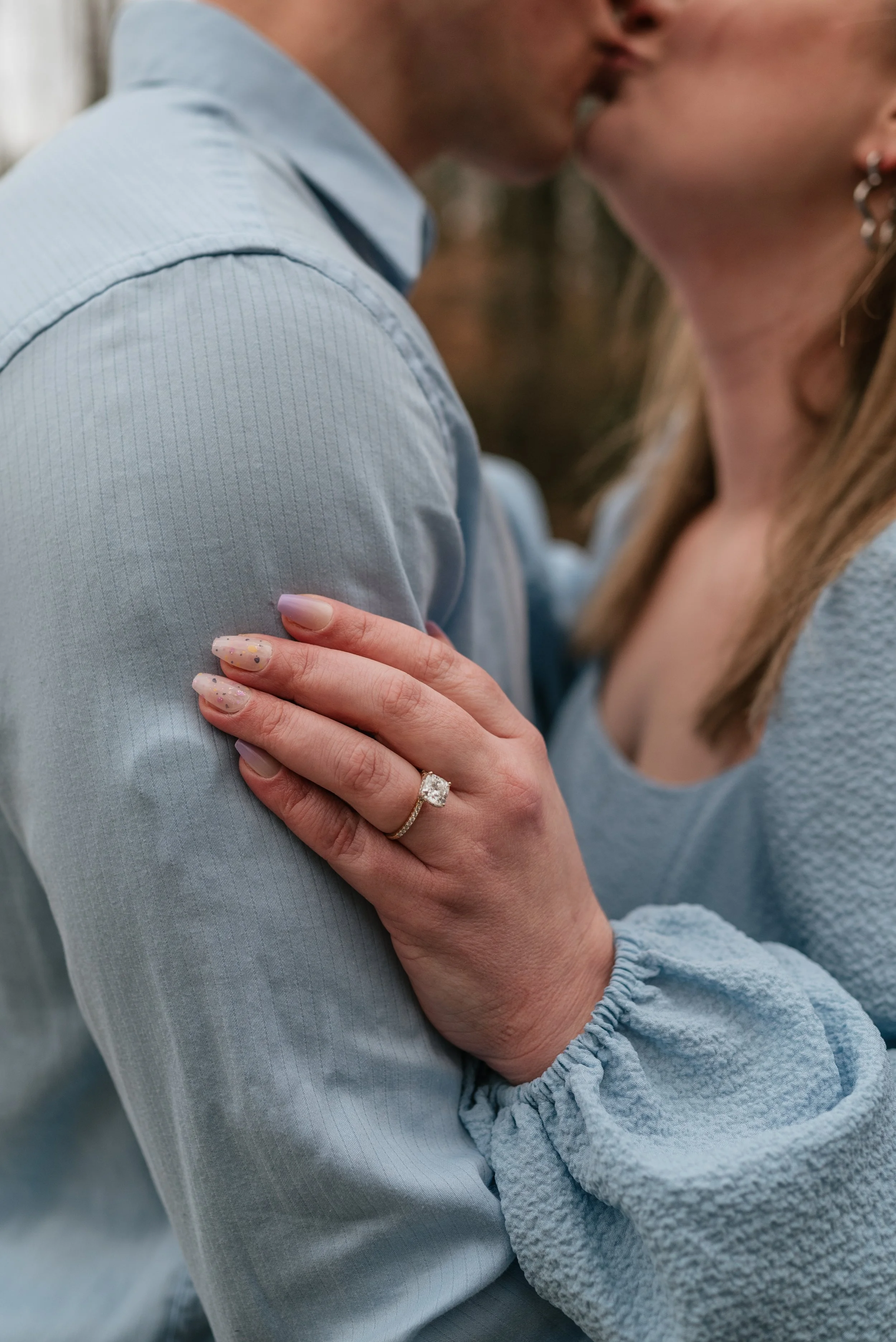 Close-up of a woman’s hand with an engagement ring resting on the shoulder of a man. They are about to kiss, dressed in light blue clothing during their engagement photoshoot in Alpharetta, GA.