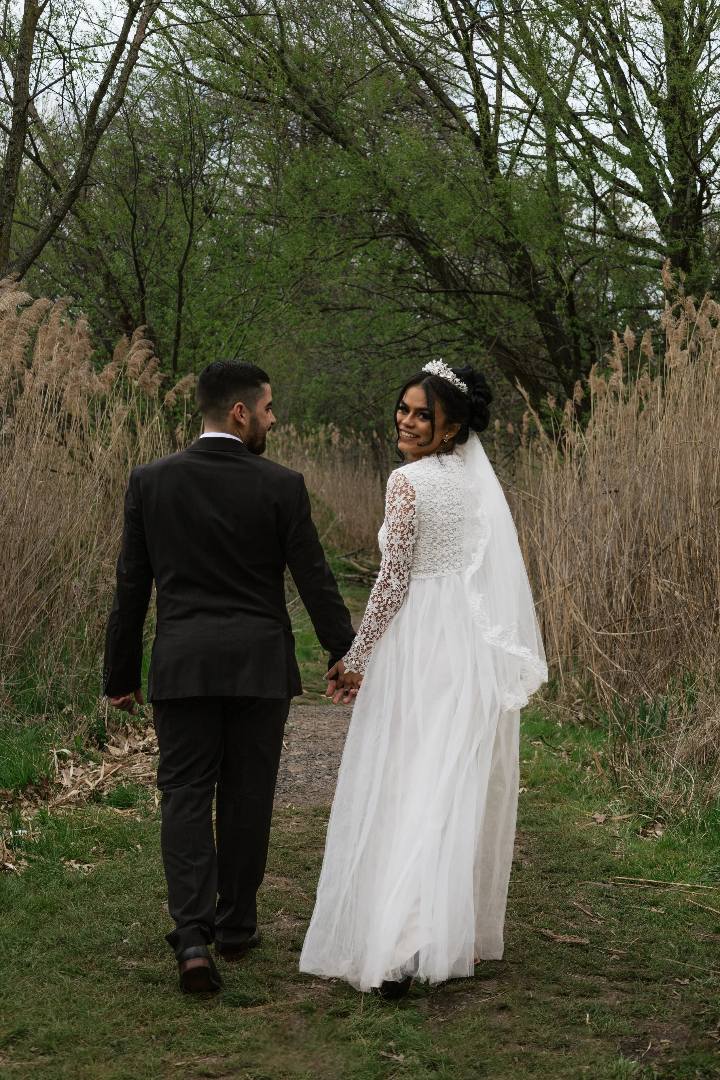 Bride and groom holding hands and walking along a nature path surrounded by trees and tall grass, bride dressed in a white lace wedding gown with a veil and tiara, groom in a black suit, smiling at each other.