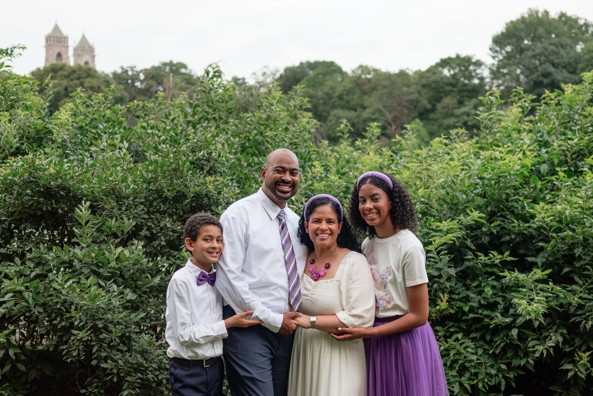 A family of four stands outdoors among greenery, smiling. The father and son are on the left, the mother and daughter are on the right. They are dressed in semi-formal clothing, with the daughter and son wearing purple accents.