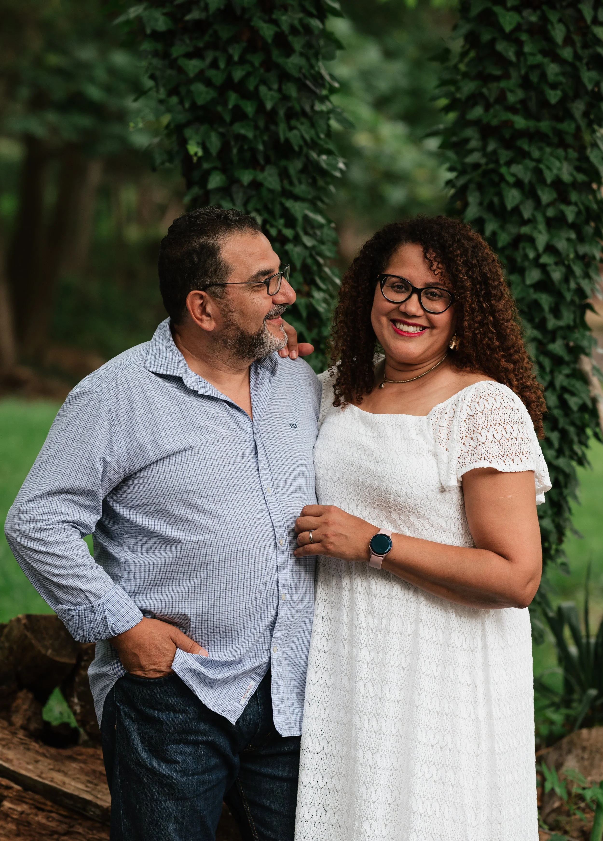 A smiling couple standing close together outdoors, with a background of green foliage and tall trees, wearing casual clothes.